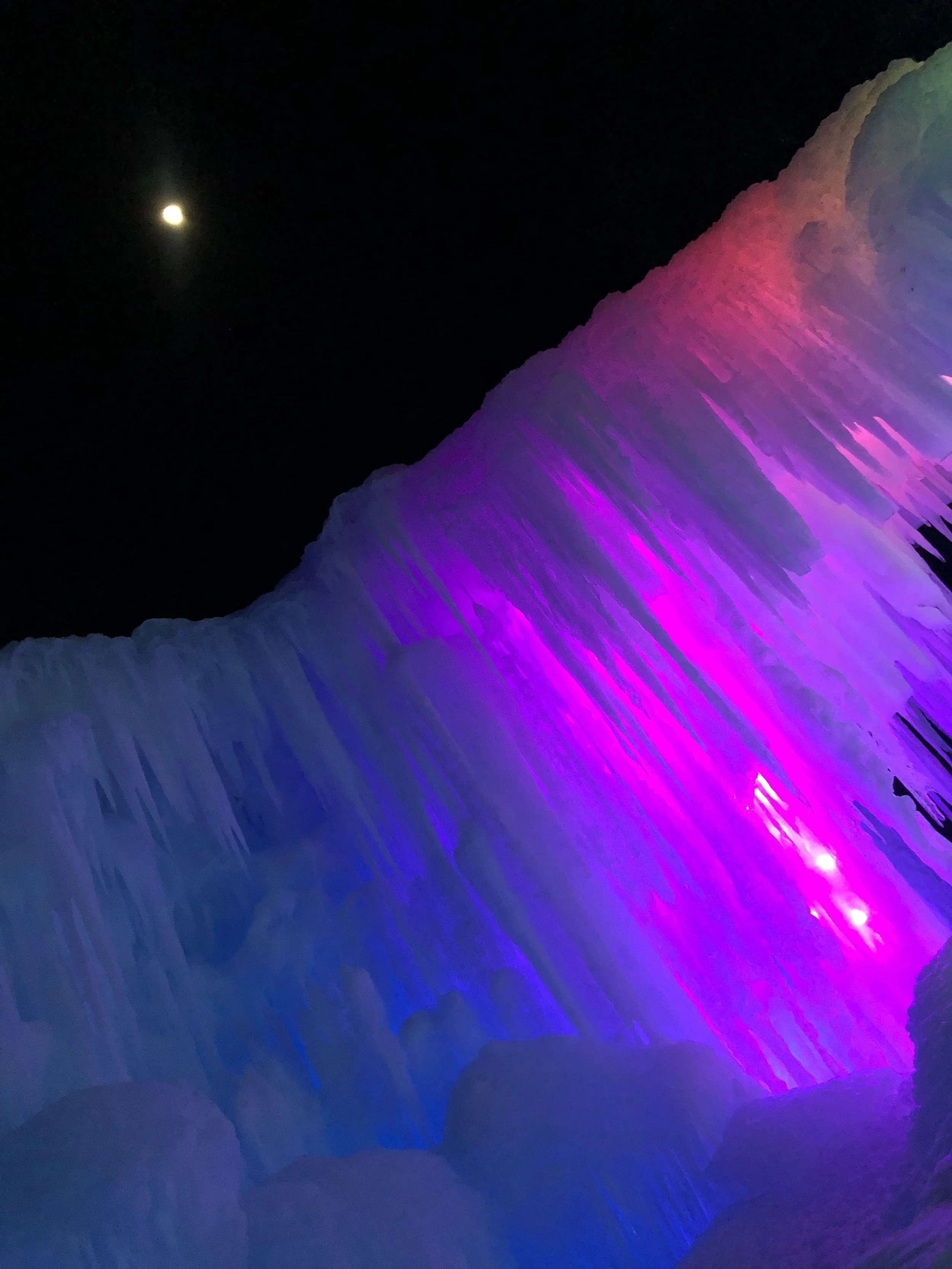 Night view of a waterfall illuminated with pink, purple, and blue lights, with a full moon in the dark sky above.