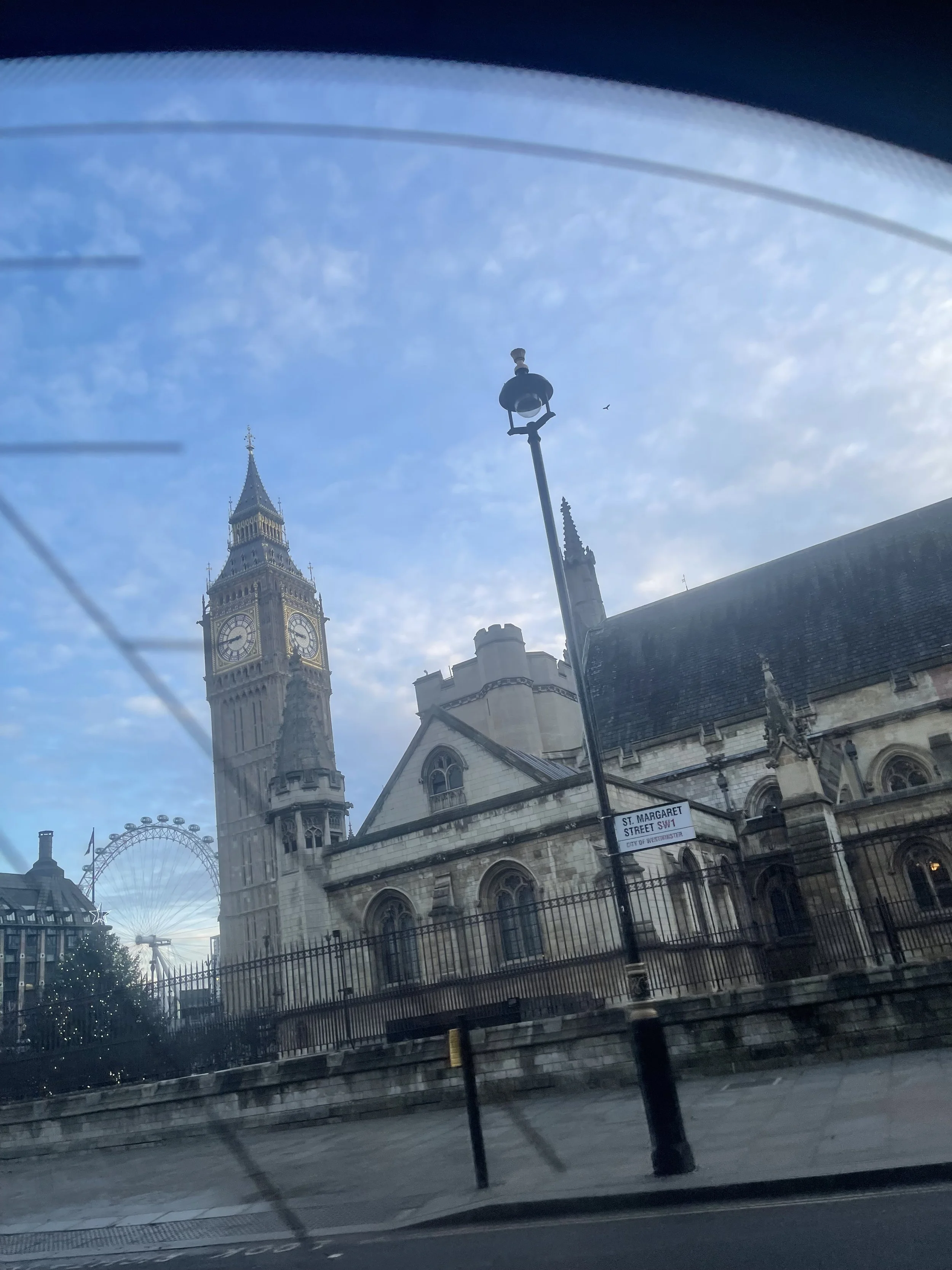 View of Big Ben, the London Eye, and a church with a spire through a window with visible wiper blades, taken from a moving vehicle.