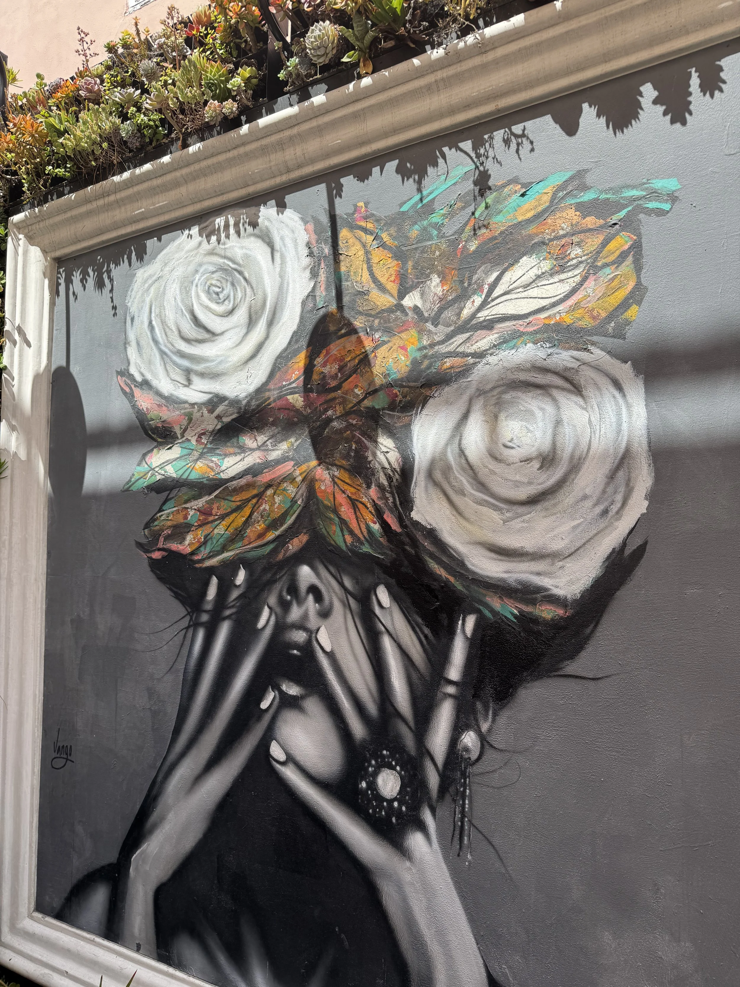 Street art mural of a woman with large, white roses as her eyes, black and white face, black hair, and wearing jewelry. Her hands are positioned near her face, and colorful leaves cover part of her head. The artwork is framed and decorated with succu