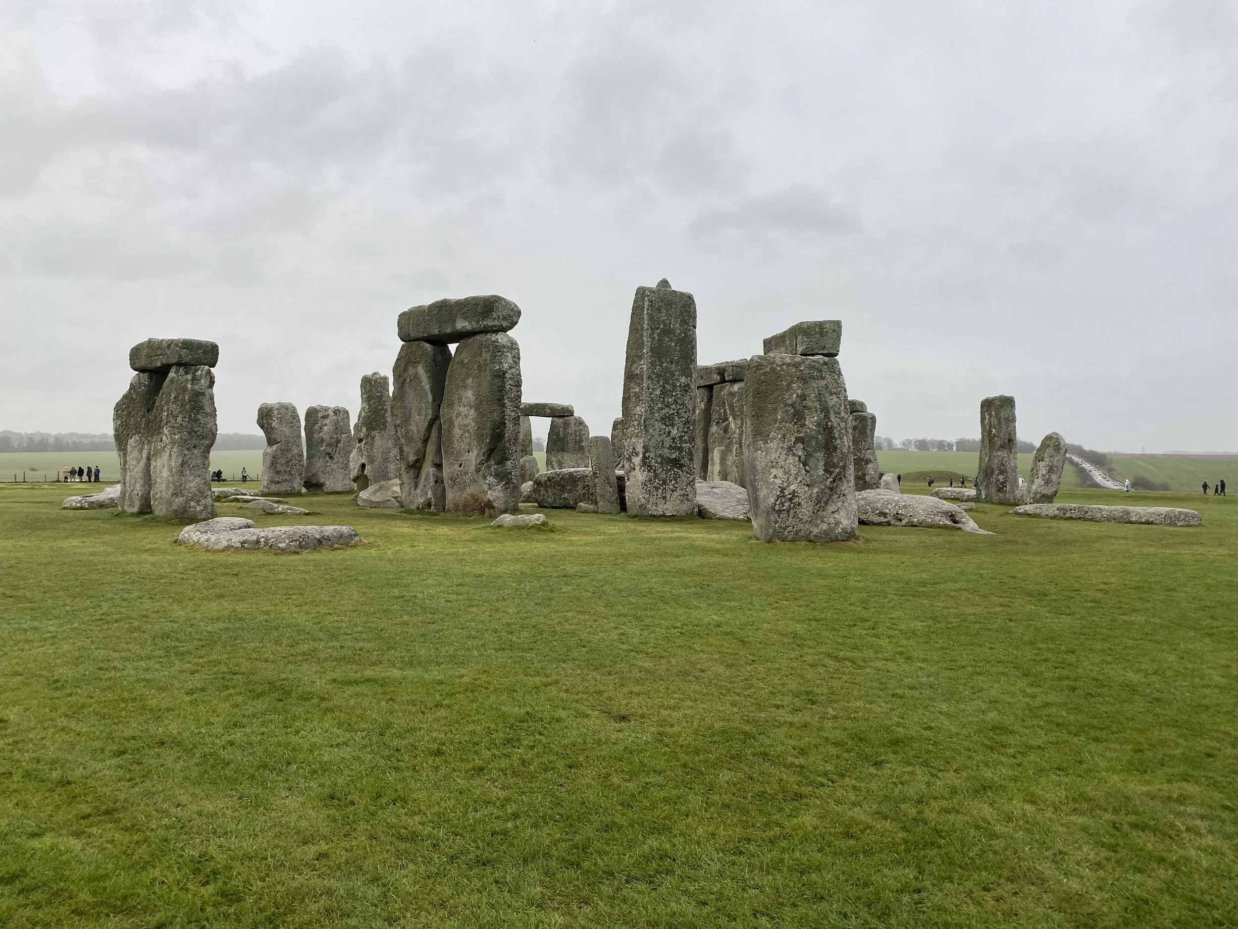 Ancient stone arrangement of standing stones at Stonehenge on a cloudy day with green grass and distant visitors.