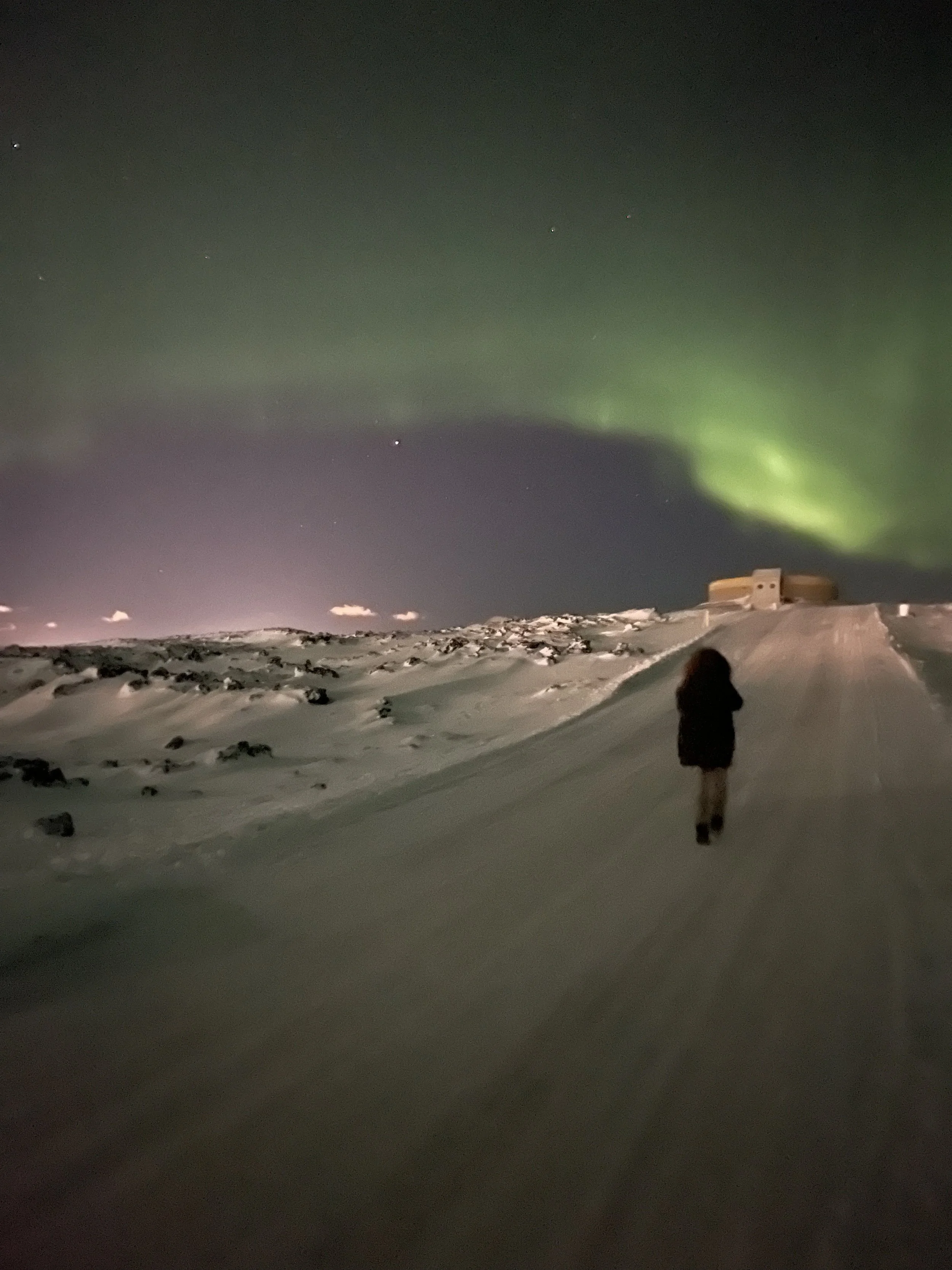 A person walking on a snow-covered road at night under the northern lights in a remote, cold landscape.