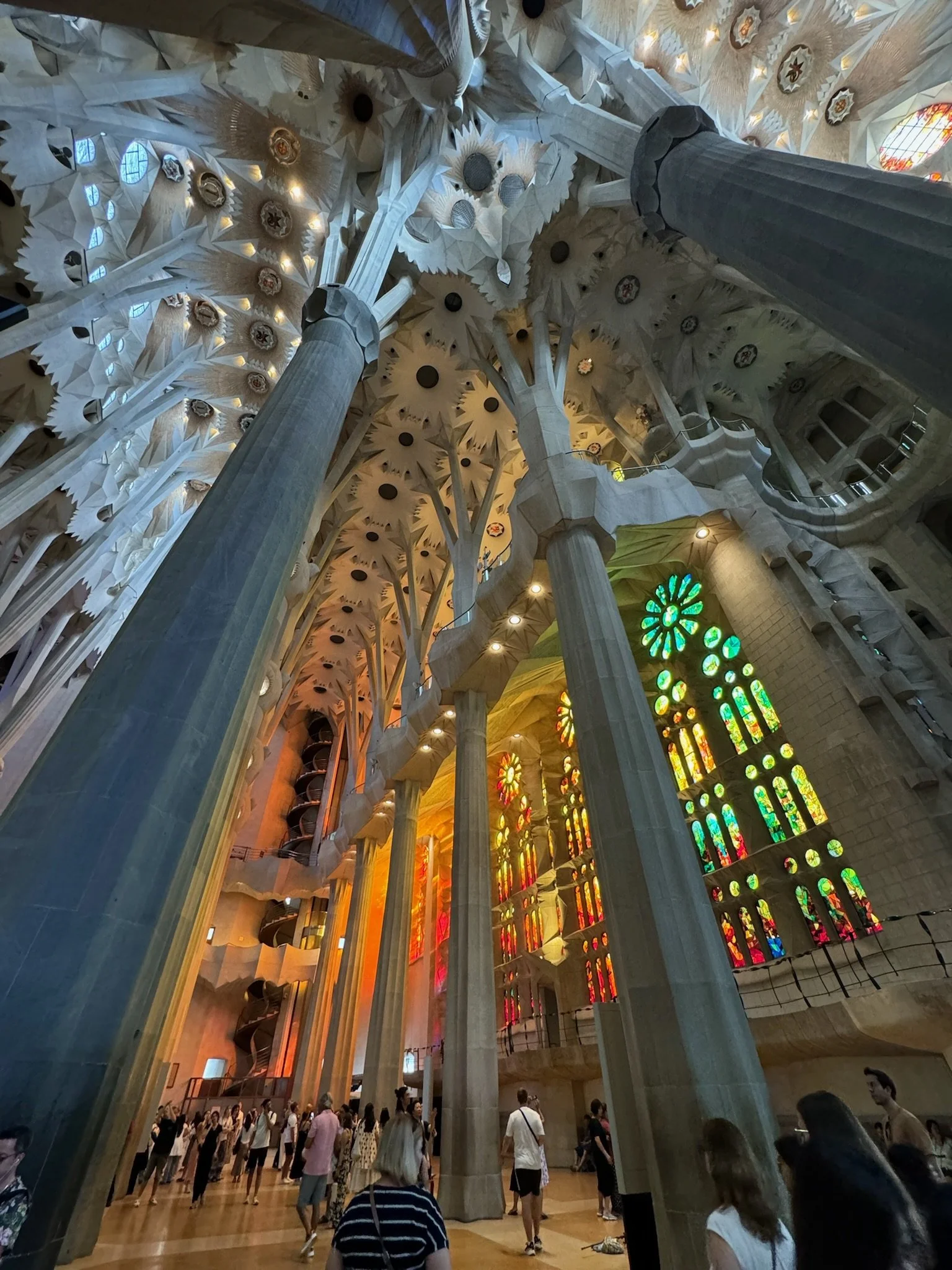 Interior of Sagrada Familia basilica showing tall columns, colorful stained glass windows, and visitors walking inside.