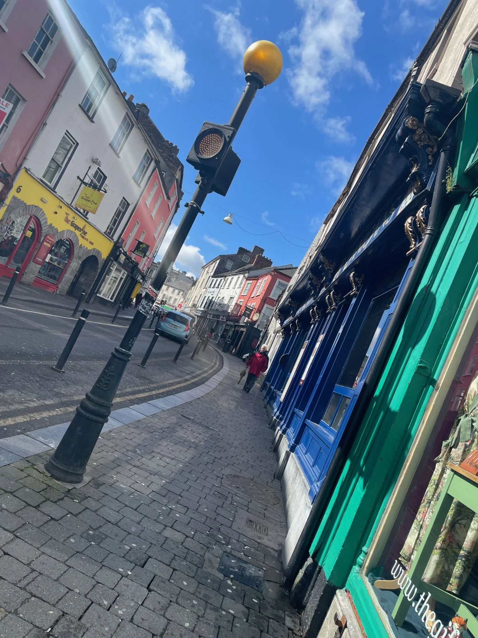 Street view with colorful buildings, a sidewalk, a traffic light pole with a yellow globe, and a person walking.