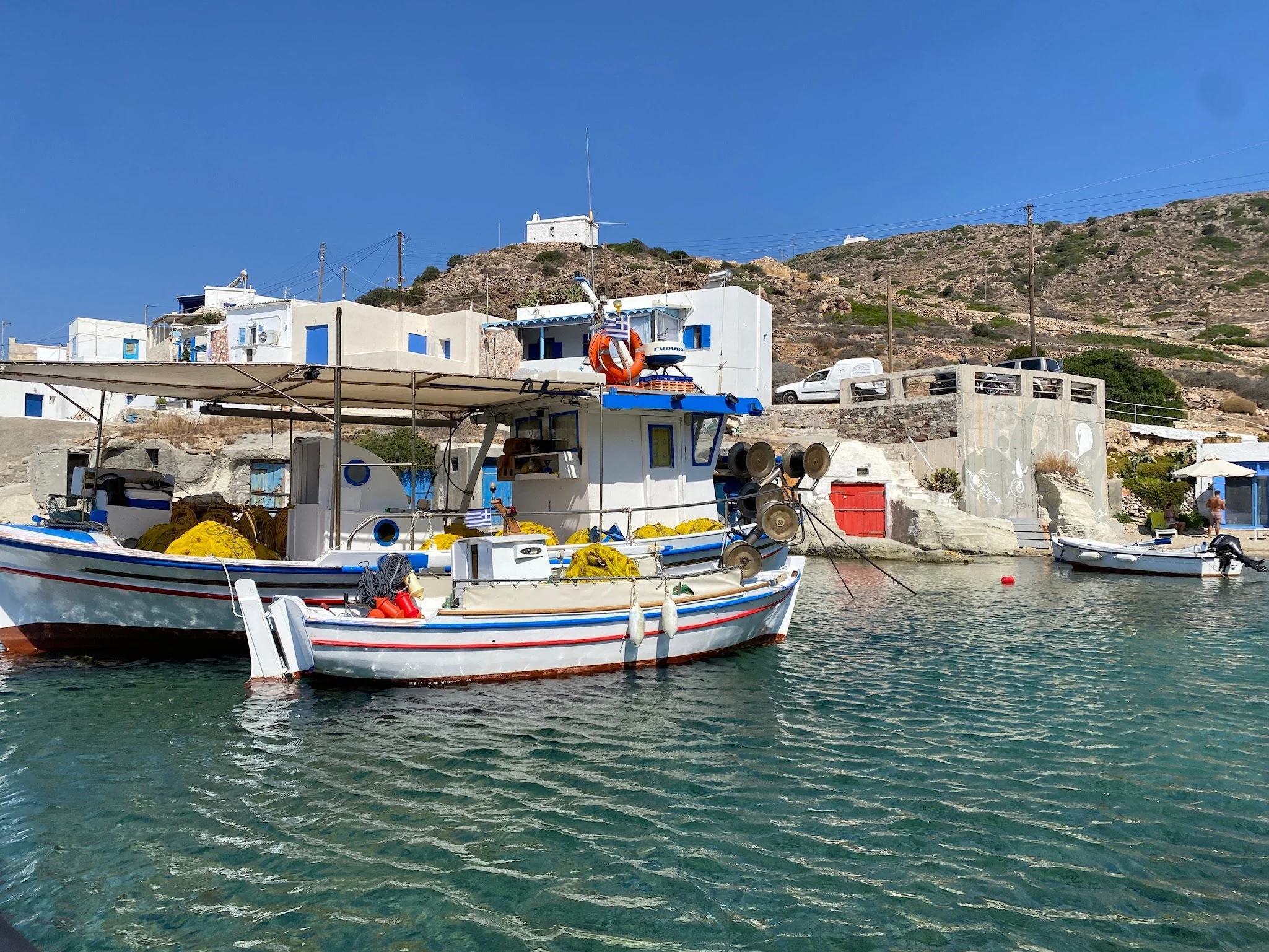 A boat in a fishing village outside of Milos, Greece docked near a harbor with buildings and a hillside in the background.