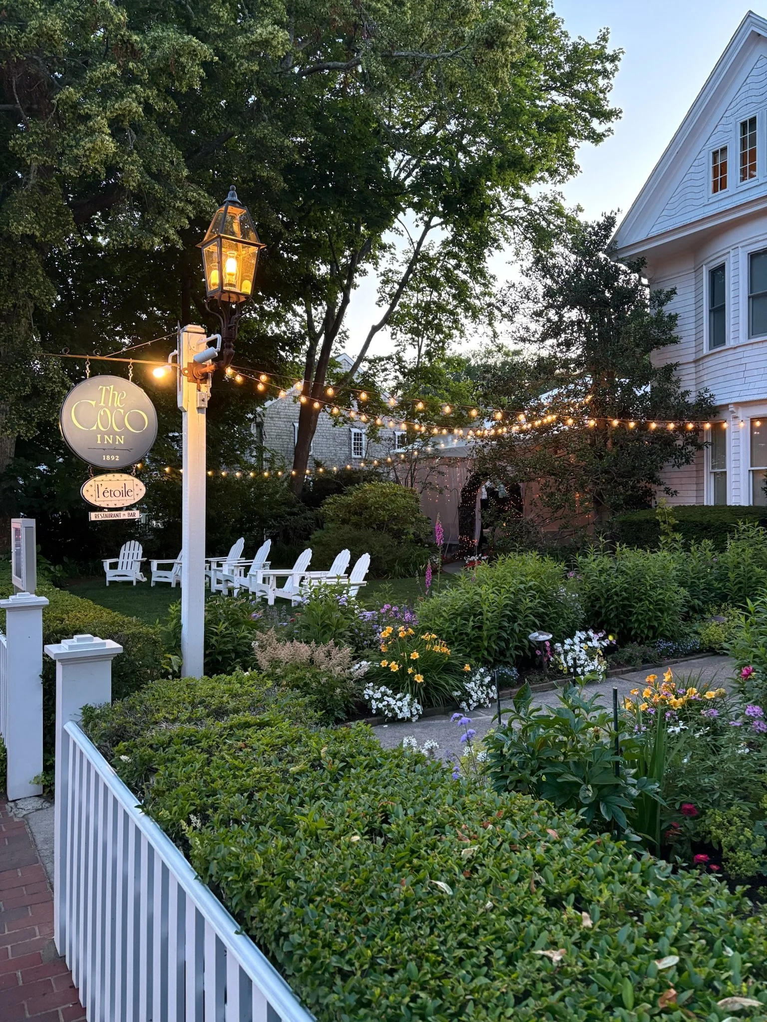 A garden scene at dusk featuring white Adirondack chairs, colorful flowers, and string lights hung across the yard, with a white house and tall trees in the background.