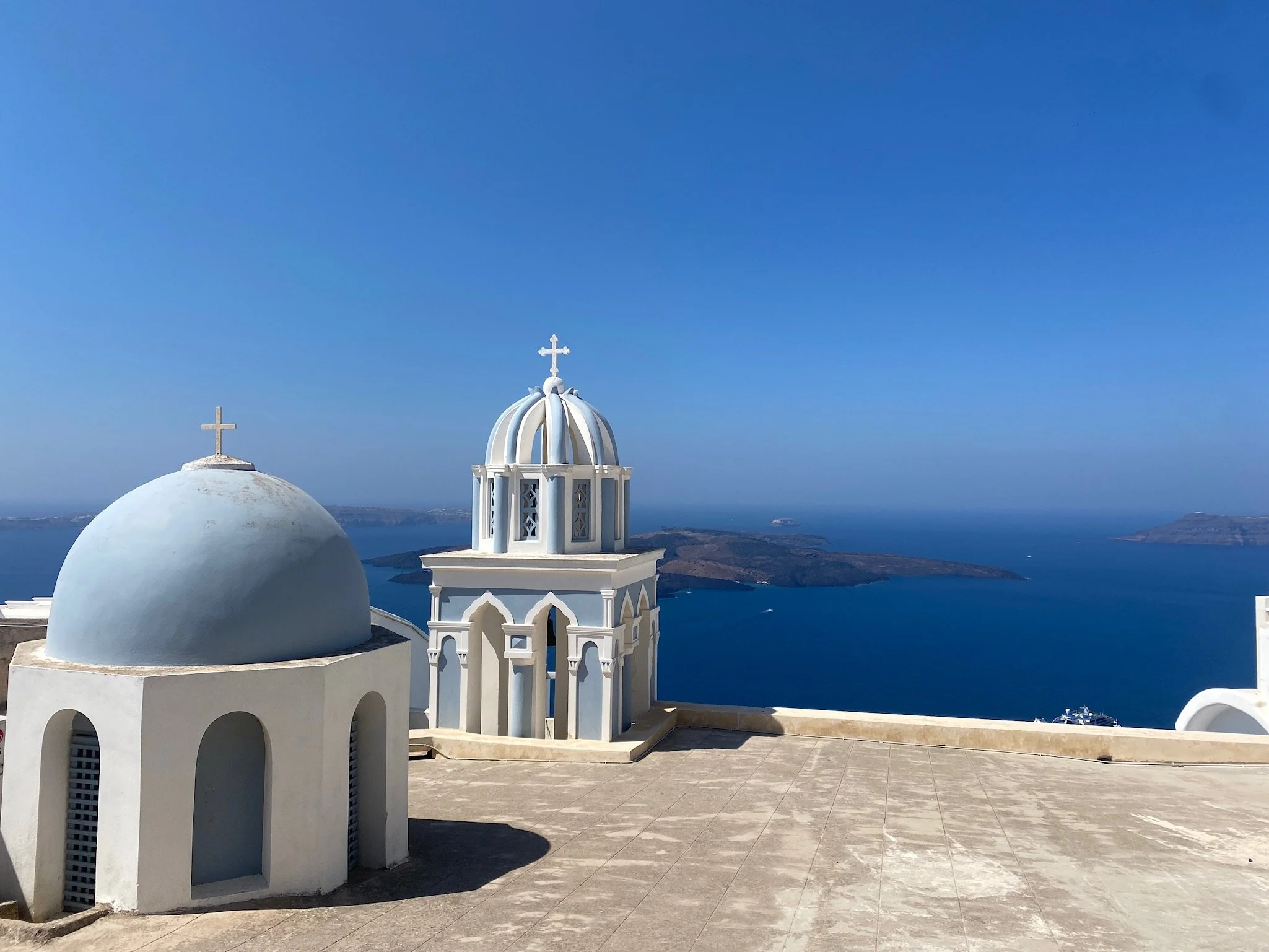White church with blue domed roof and bell tower on a terrace overlooking the sea and islands.