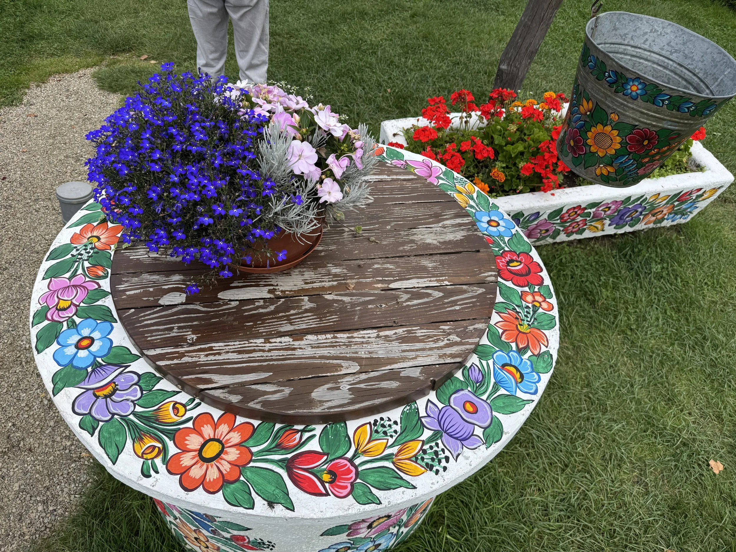 Colorful flower pots with vibrant blooms, including purple, pink, red, and orange flowers, on a decorated round wooden table and a rectangular planter, outdoors on grass and gravel.