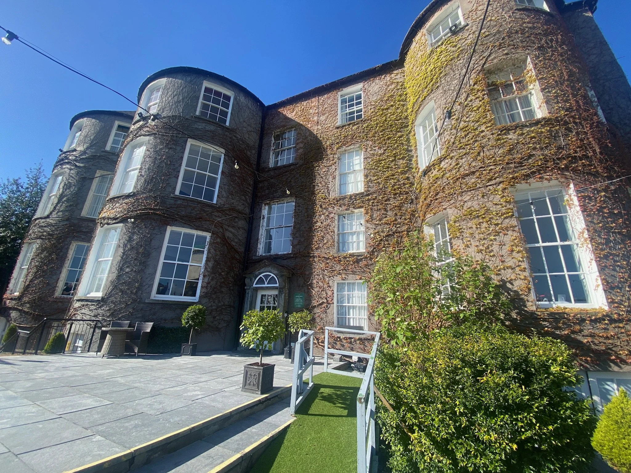 A multi-story building with arched windows, covered in climbing vines, with a cleared paved front area and green bushes along the side, under a clear blue sky.