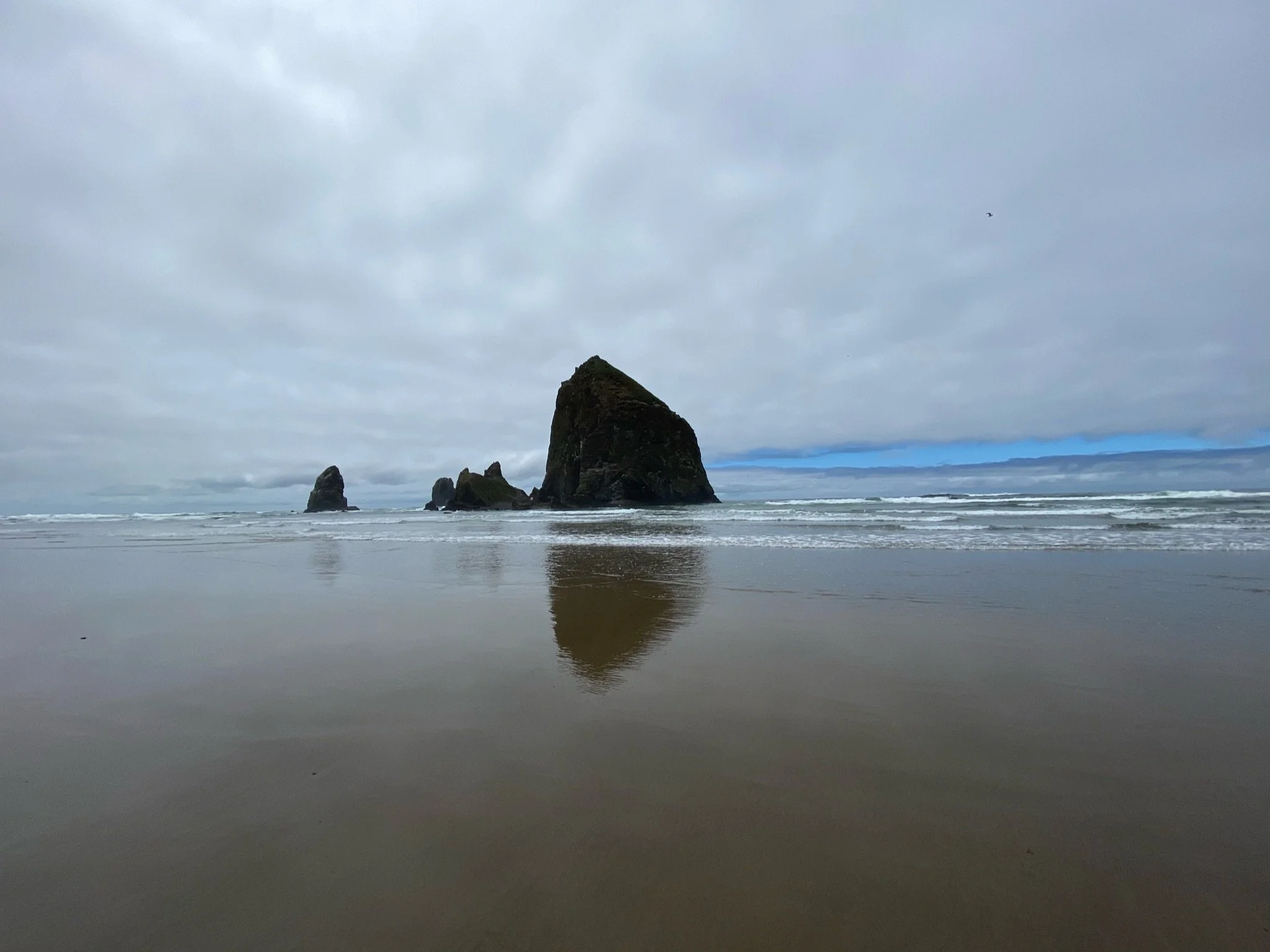Overcast beach with large sea stacks, including Haystack Rock, reflected in wet sand, with waves and cloudy sky in the background.