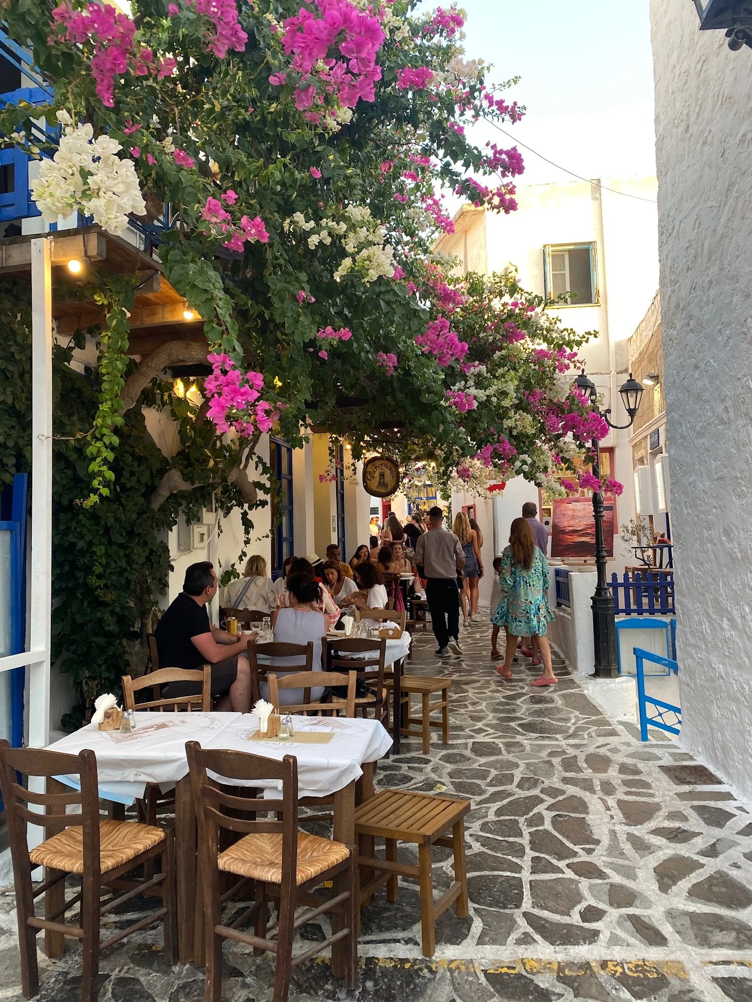 Outdoor restaurant with tables covered in white tablecloths, wooden chairs, and blooming pink and white bougainvillea on a narrow cobblestone street. People are dining and walking under the flowers near white buildings with blue accents, typical of a