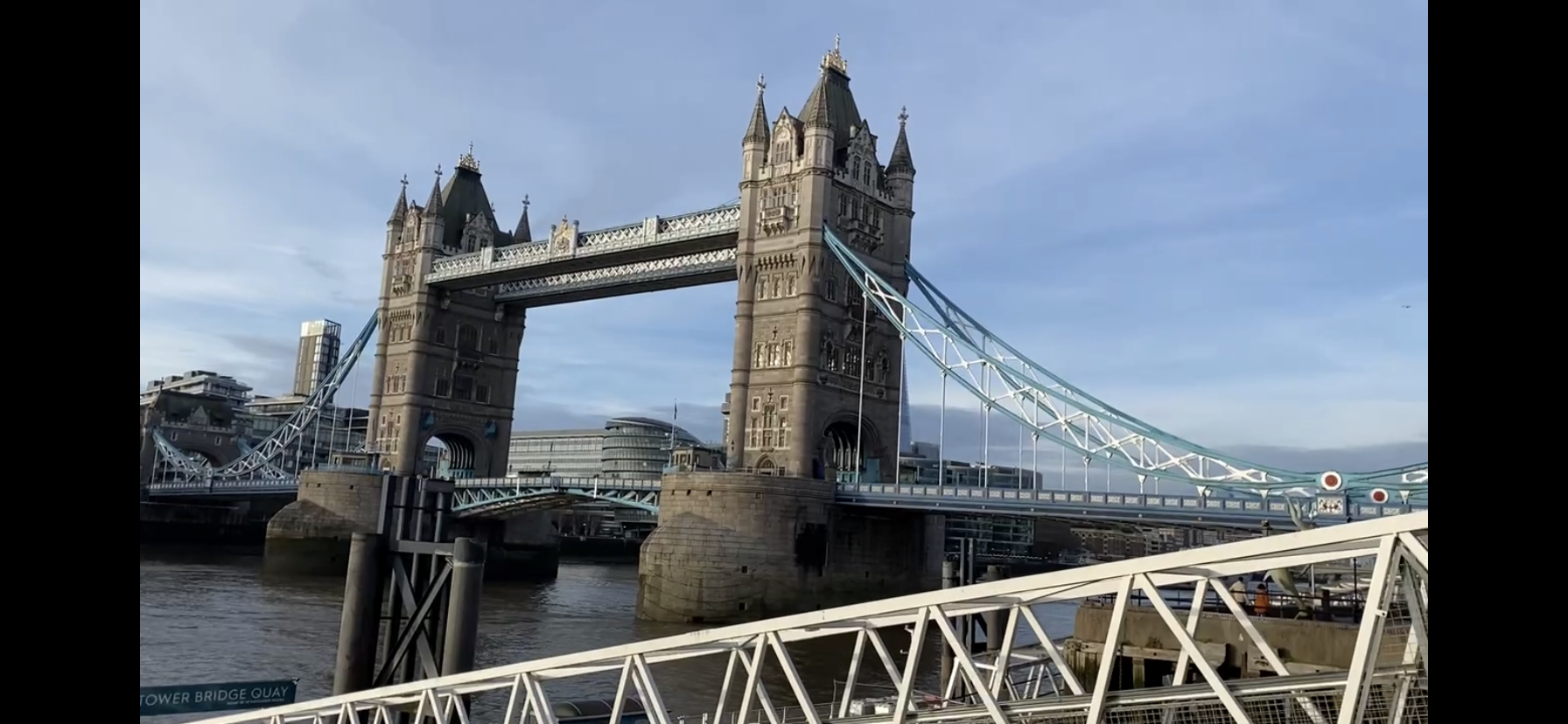 View of Tower Bridge in London with the River Thames in the foreground and modern buildings in the background, under a clear blue sky.