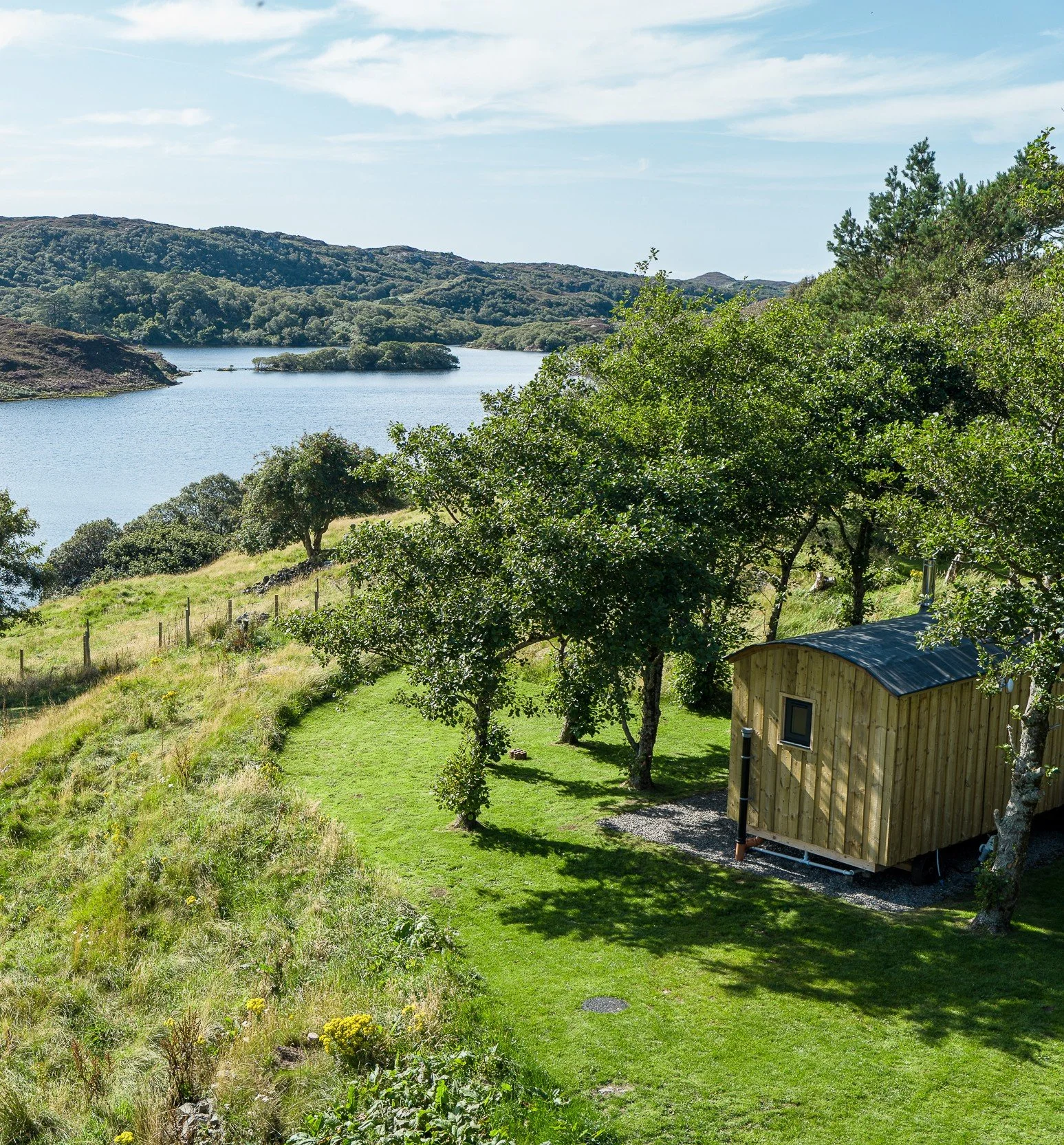 A scenic view of Loch Drumbeg surrounded by lush green trees and rolling hills, with a shepherd's hut in the foreground.