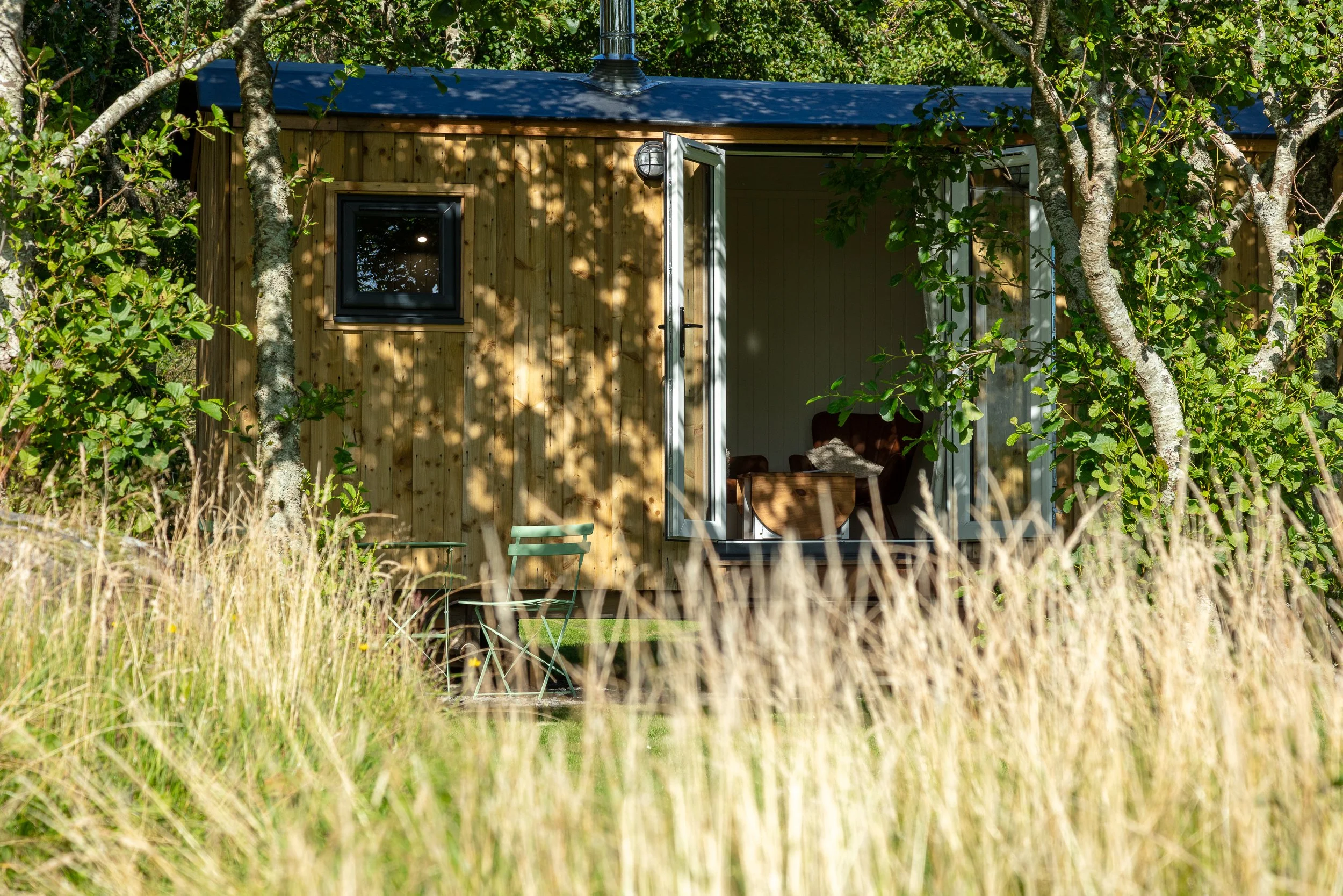 A shepherd's hut surrounded by trees with an open French door revealing a cosy interior with chairs and a table.