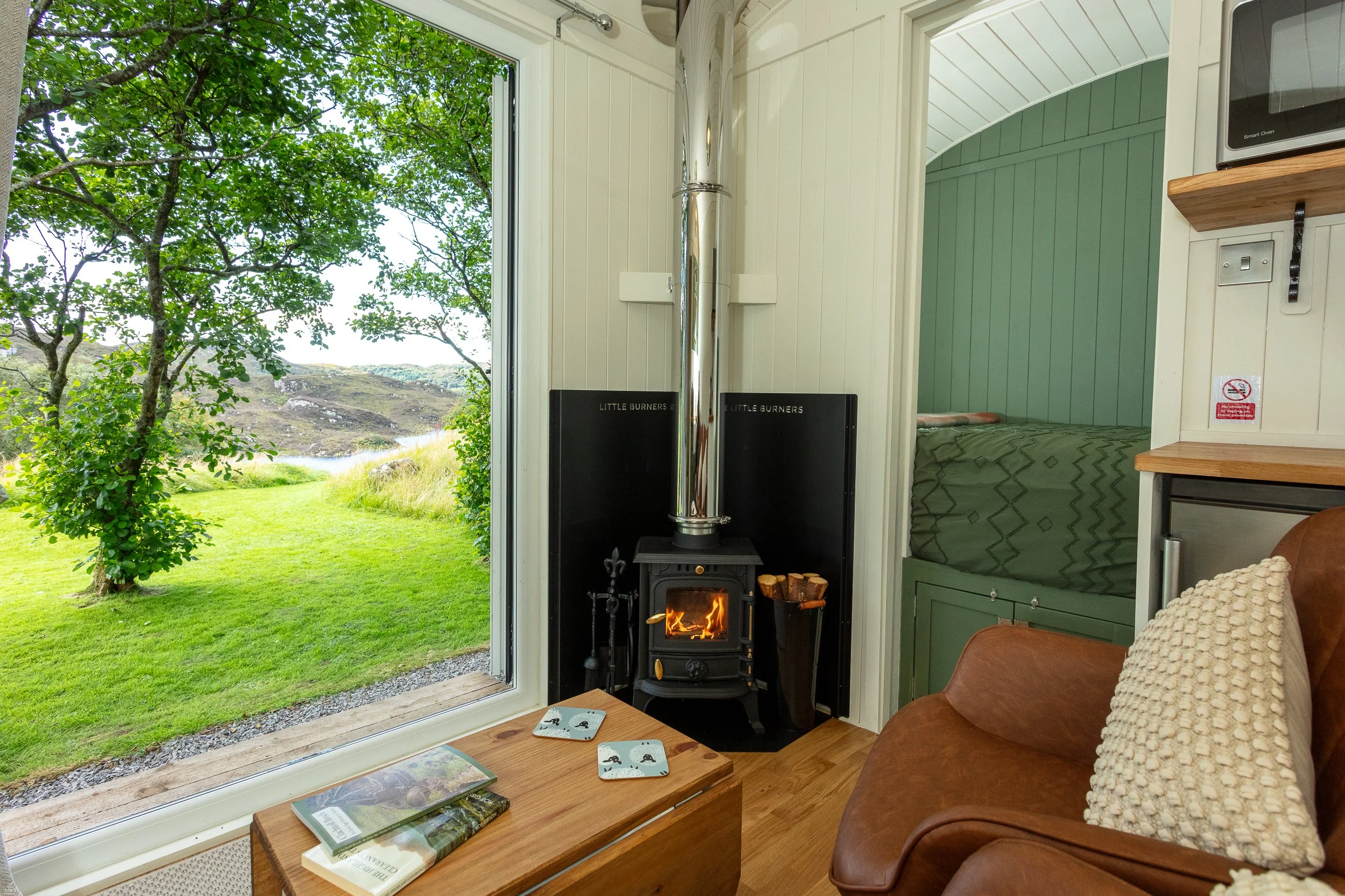 Interior of a cosy shepherd's hut with a wood stove burning and a view of green trees and Loch Drumbeg through the open French doors.