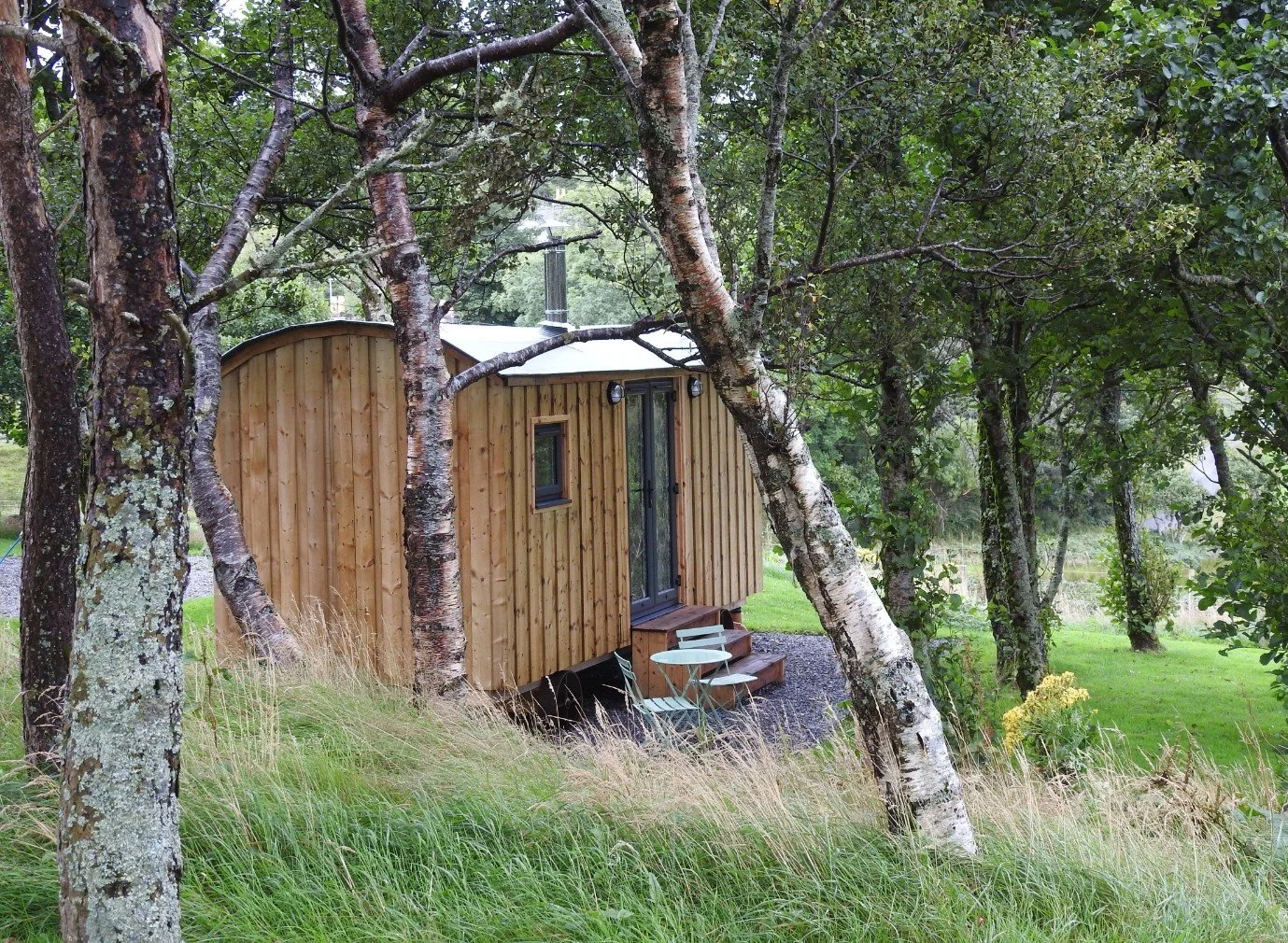 A shepherd's hut nestled among trees with steps leading to a French door, outdoor chairs, and lush greenery.
