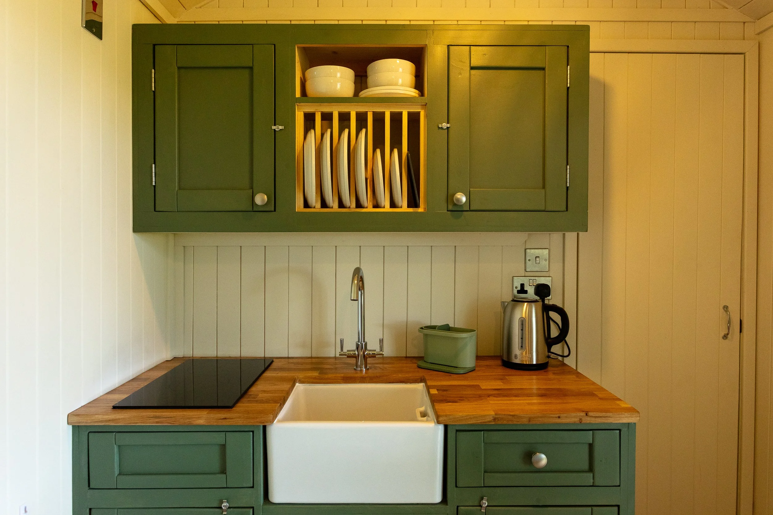 Rustic dresser-style green kitchenette with open shelf holding white bowls and plates, above a wooden countertop with a sink, a green container, a silver electric kettle, and a ceramic hob.