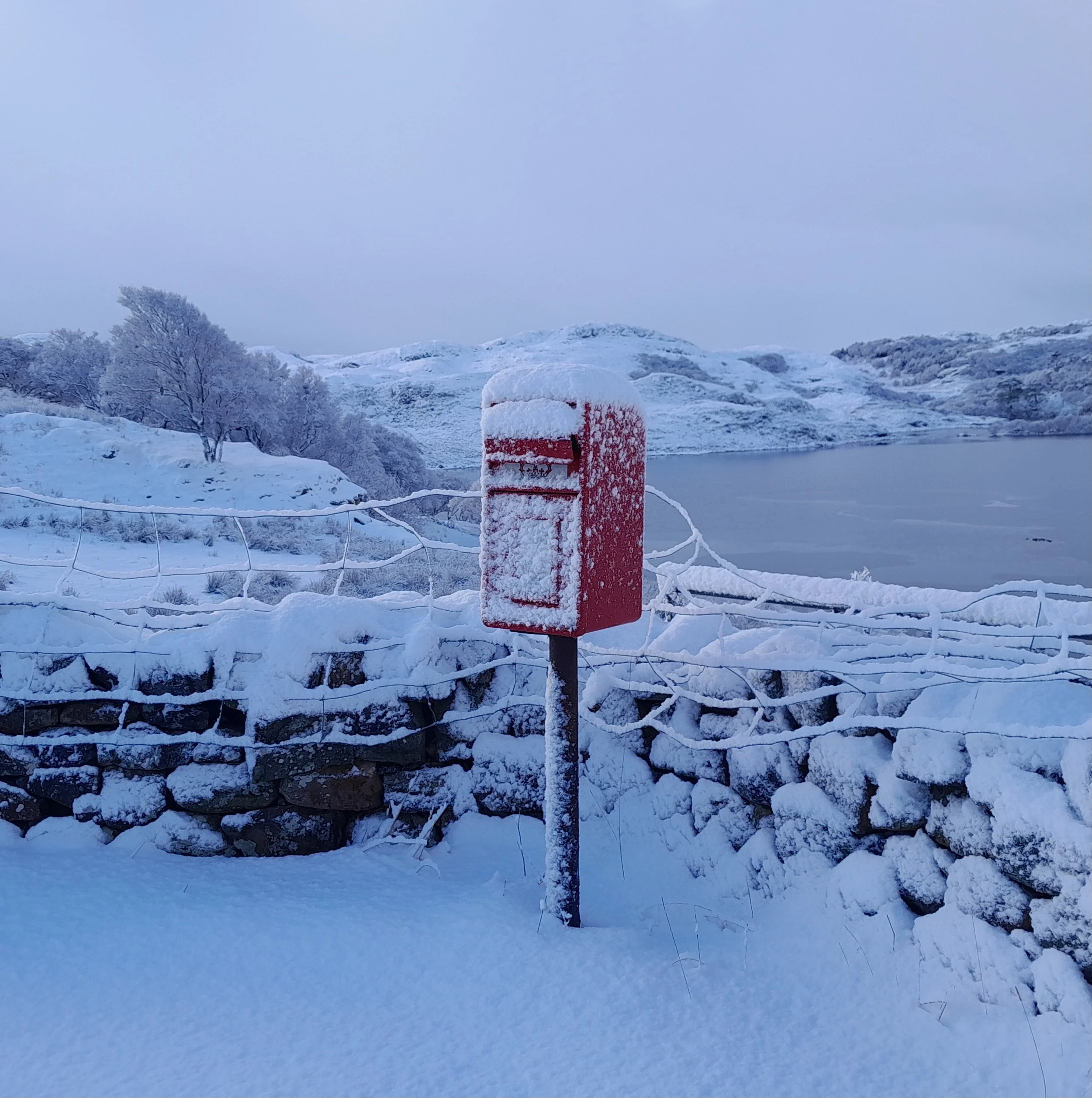 Snow-covered landscape with distant hills and bare trees, a frozen Loch Drumbeg, and a snow-covered postbox in the foreground.