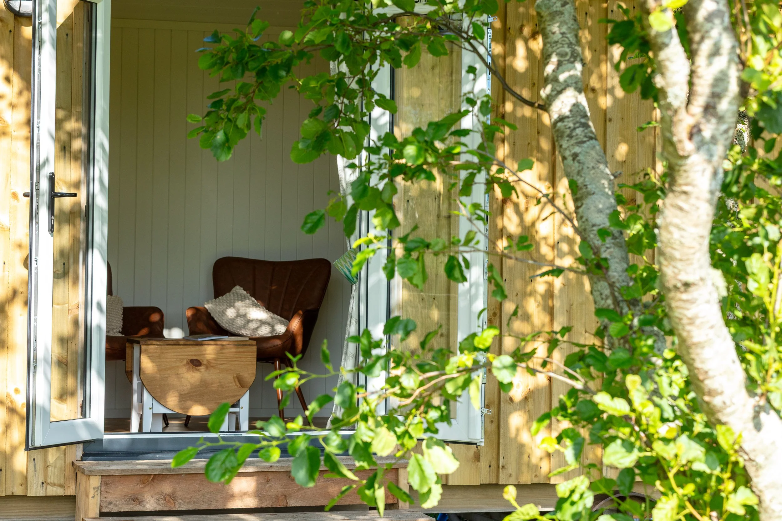 Interior view of a cosy room with two brown armchairs, a wooden coffee table, and white paneled walls, seen through an open door with a wooden frame, surrounded by green tree leaves.