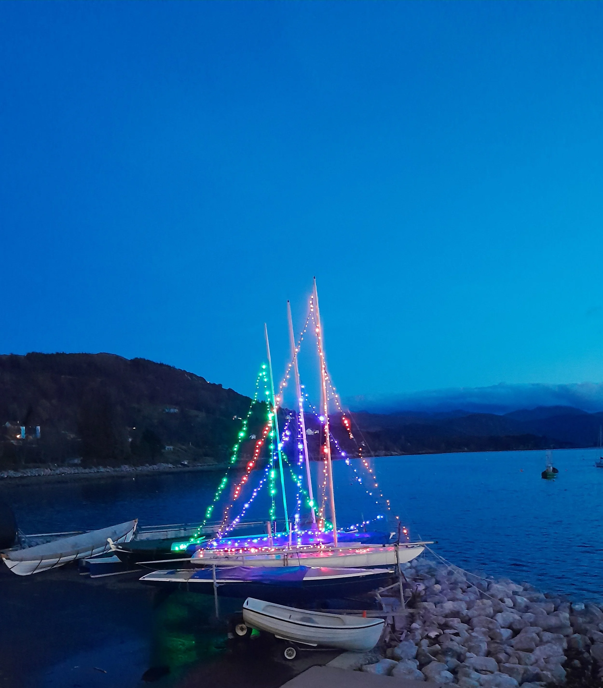 Boats docked by the Ullapool shoreline at night, with a large yacht decorated with colourful Christmas lights, set against the sky at dusk and a hill in the background.