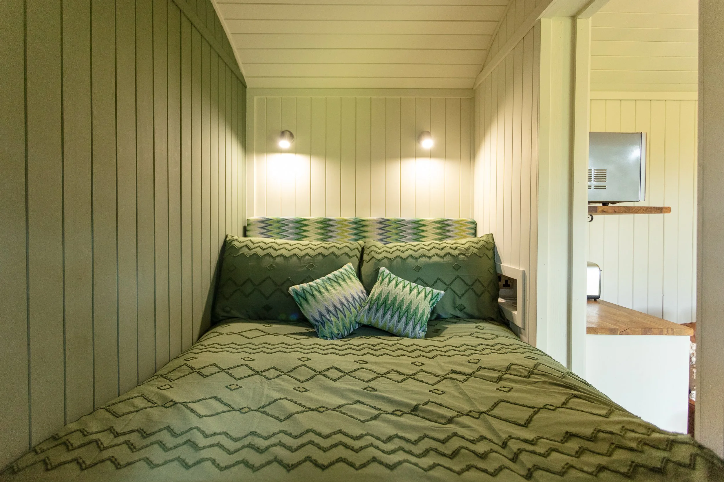 Interior of a shepherd's hut sleeping nook with light green and white panelled walls, featuring a bed with a patterned bedspread and matching pillows, two wall-mounted lights, and an open doorway revealing the living area beyond.