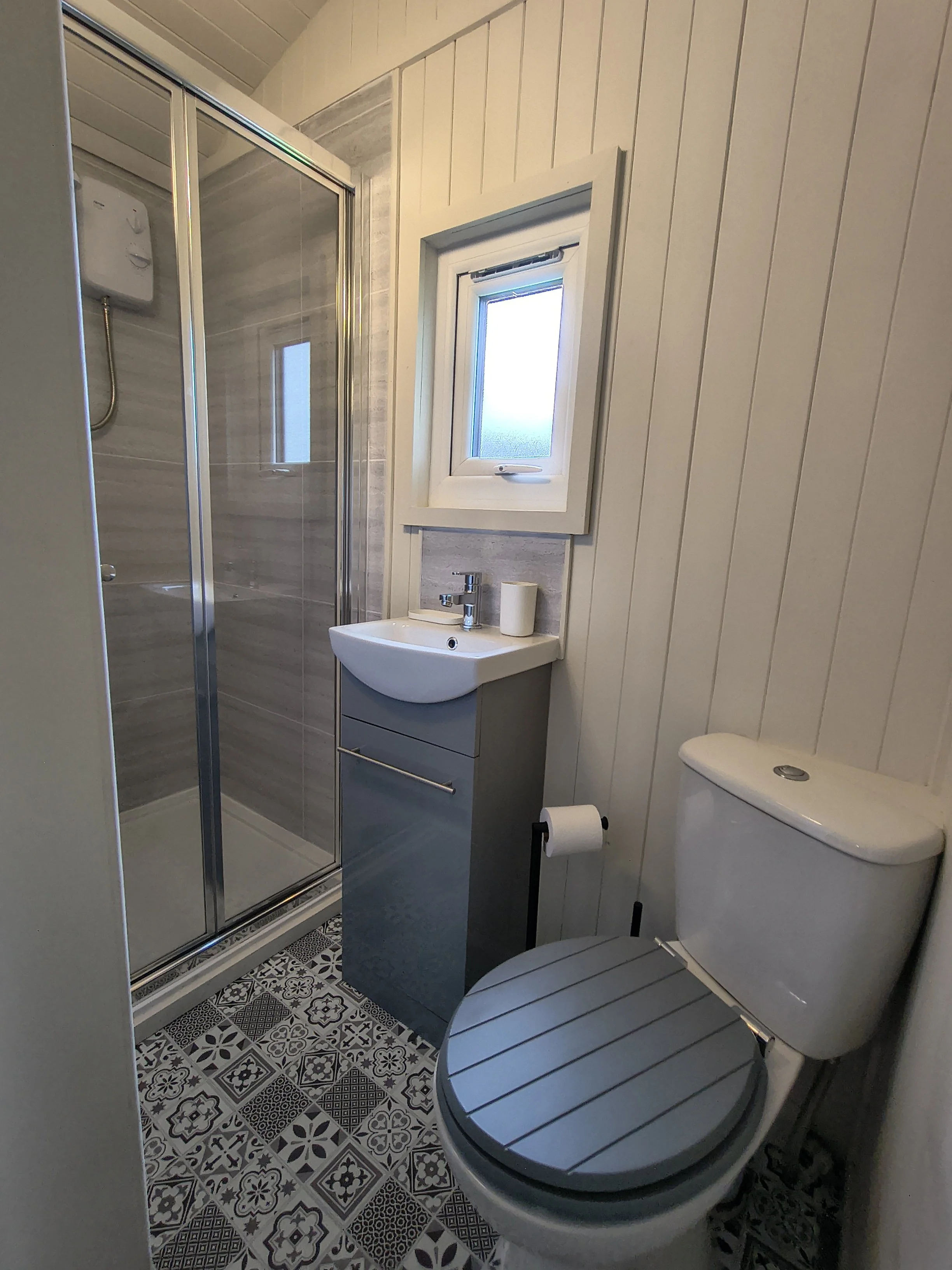 A shower room with shower enclosure, basin with cupboard beneath, a toilet with a wooden seat, patterned grey and white floor tiles, and a small window with frosted glass.