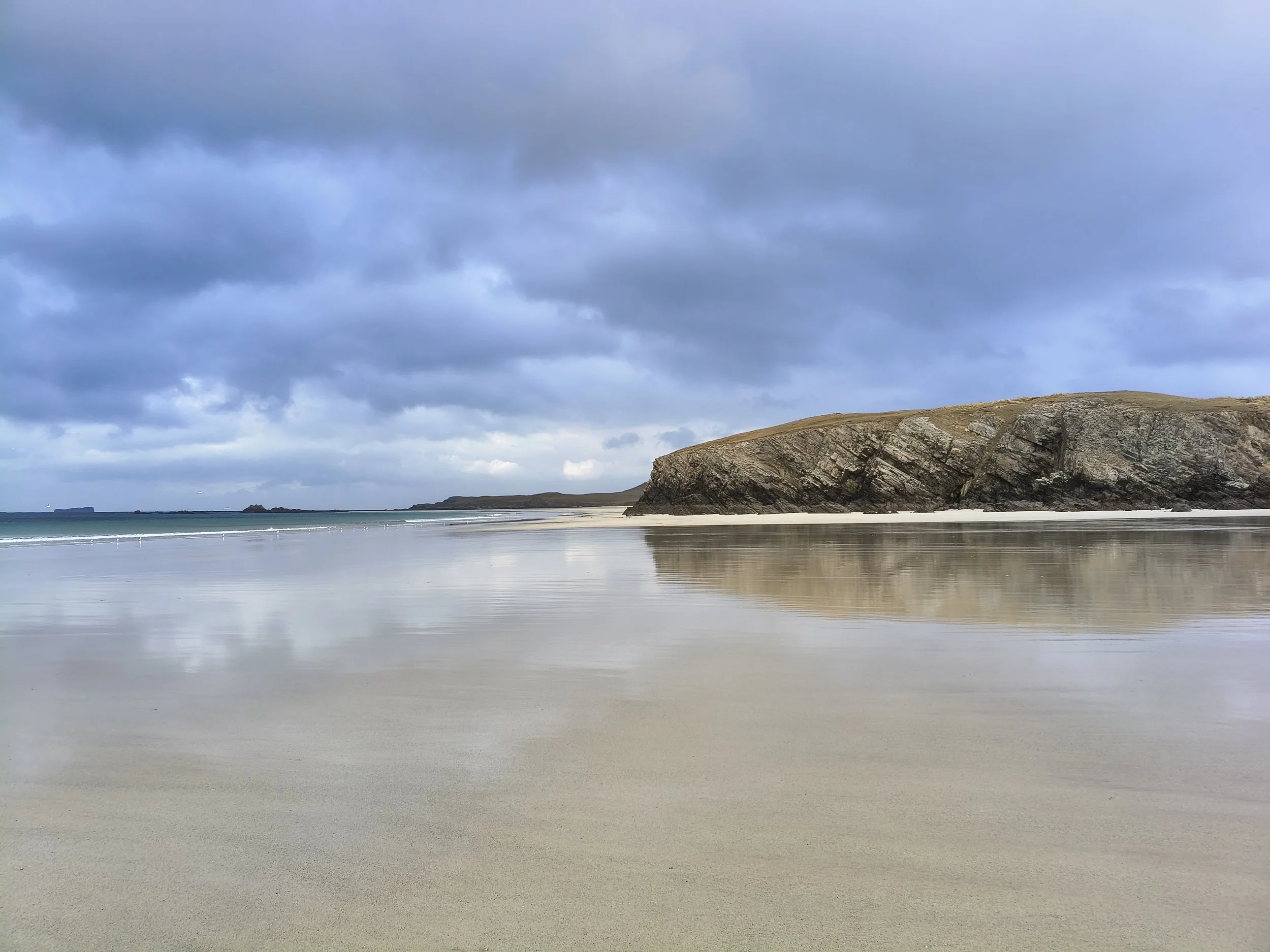 A wide sandy beach at Balnakeil, with a large rocky cliff on the right and calm ocean waters under a cloudy sky.