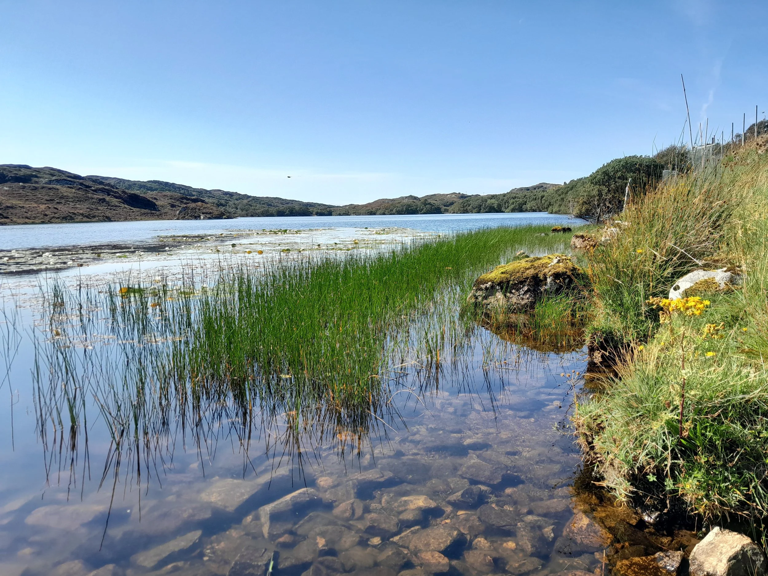 A scenic landscape of Loch Drumbeg with partially frozen water, surrounded by rocky shores and hills with sparse vegetation, under a partly cloudy sky.