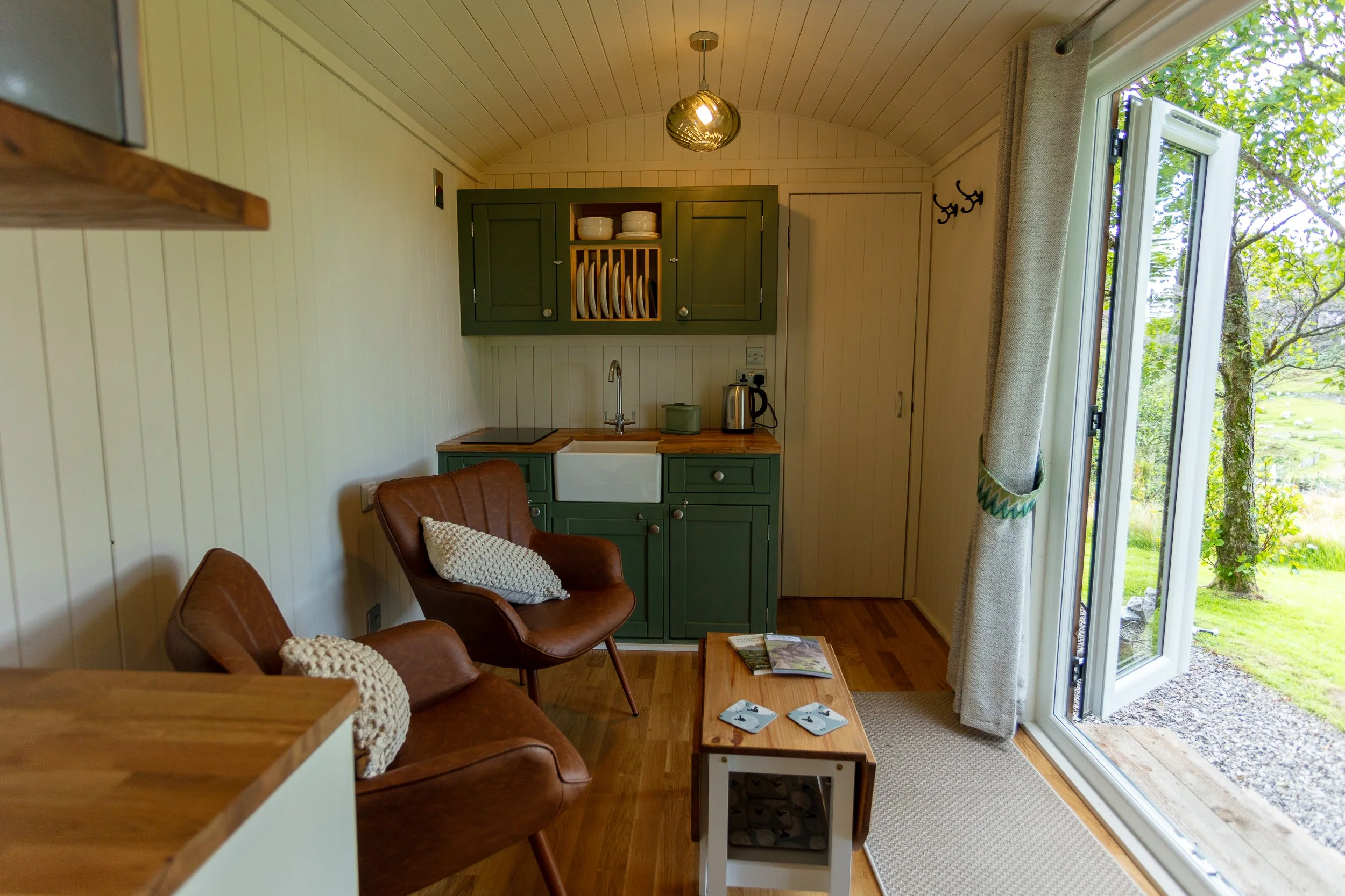 The main living area of a shepherd's hut, showing a kitchenette, two large armchairs and a table, and the doorway to the shower room, next to open French doors showing a green outdoor landscape with trees and grass.