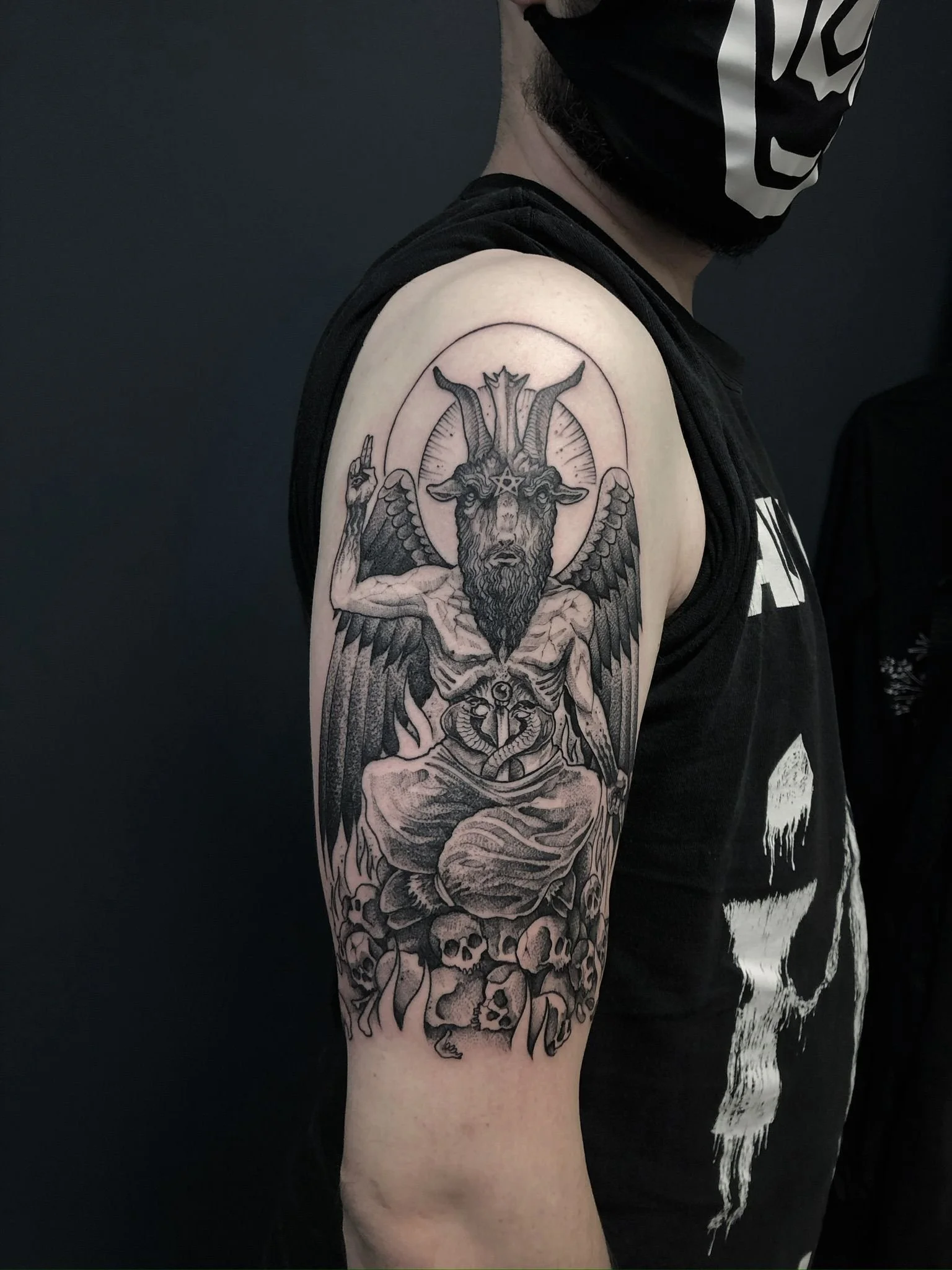 A black and gray tattoo on a man's upper arm featuring a mythological creature with a goat's head, wings, and a human body, sitting on a pile of skulls and bones, with a halo-like circle behind its head.