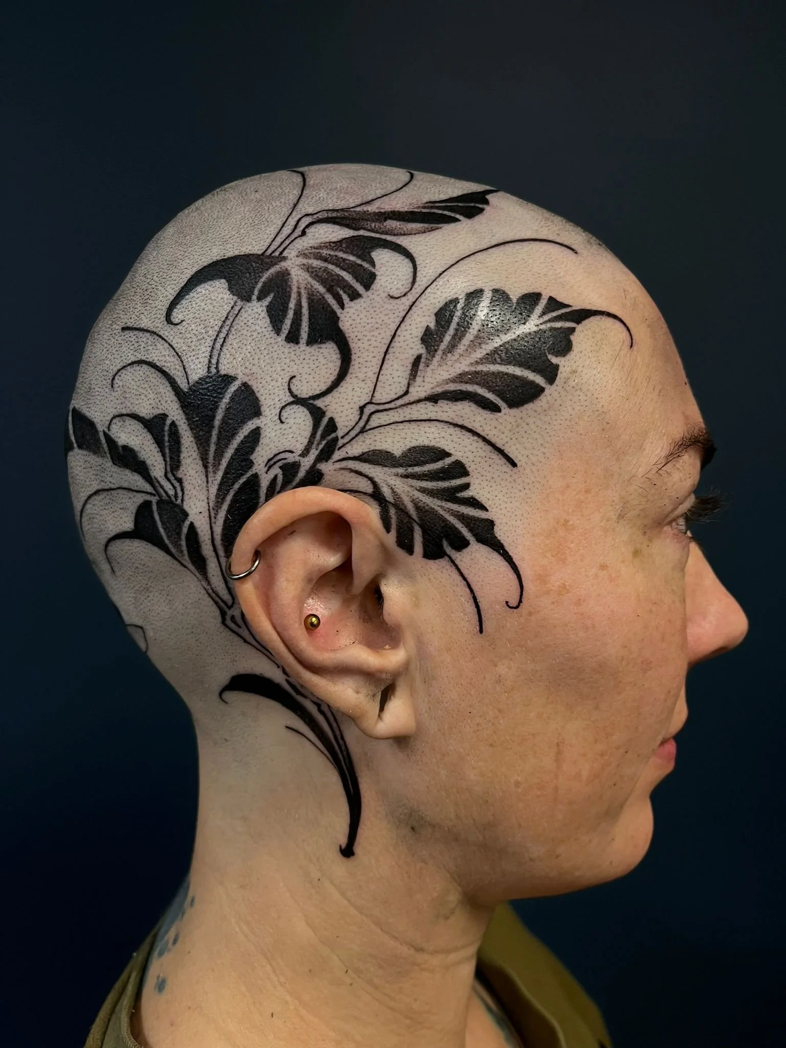 Side profile of a person with a large black floral tattoo on their shaved scalp, multiple ear piercings, and light skin.