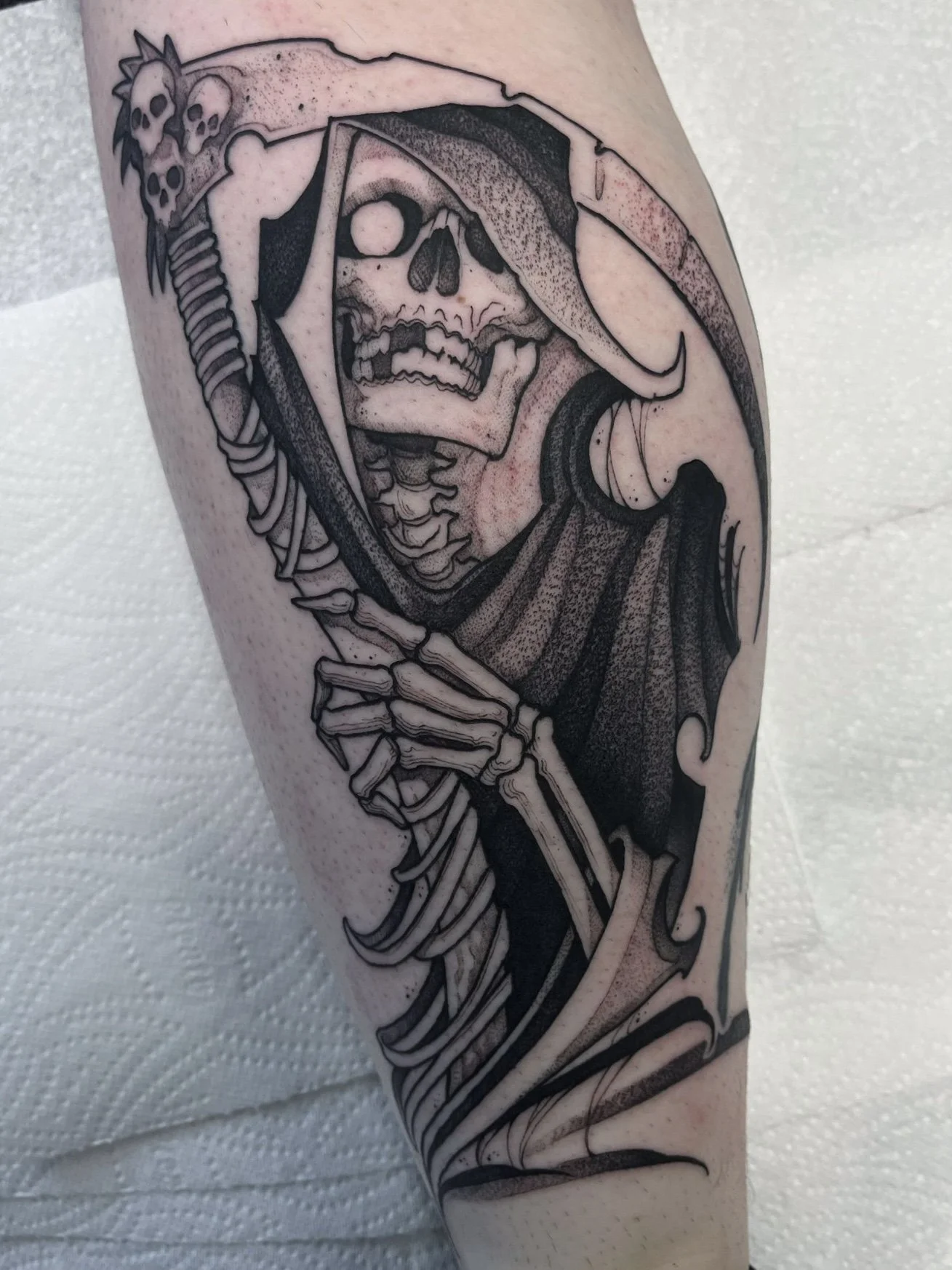 Black and gray tattoo of a skeleton with an opening skull and a grin, wearing a hood and holding a scythe, on a person's arm.