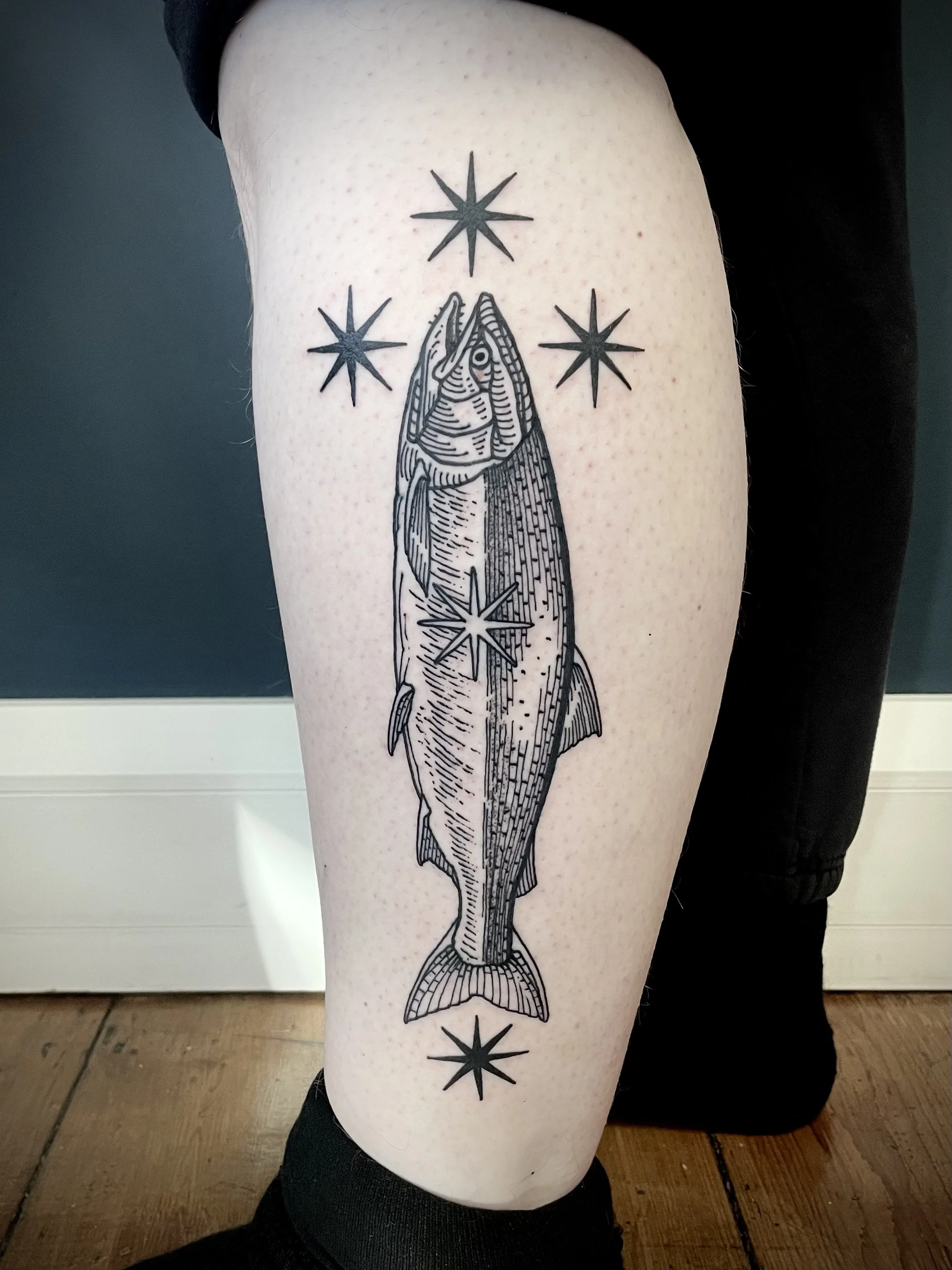 A black line tattoo of a fish with a stylized design, surrounded by four star-like shapes on a person's calf.