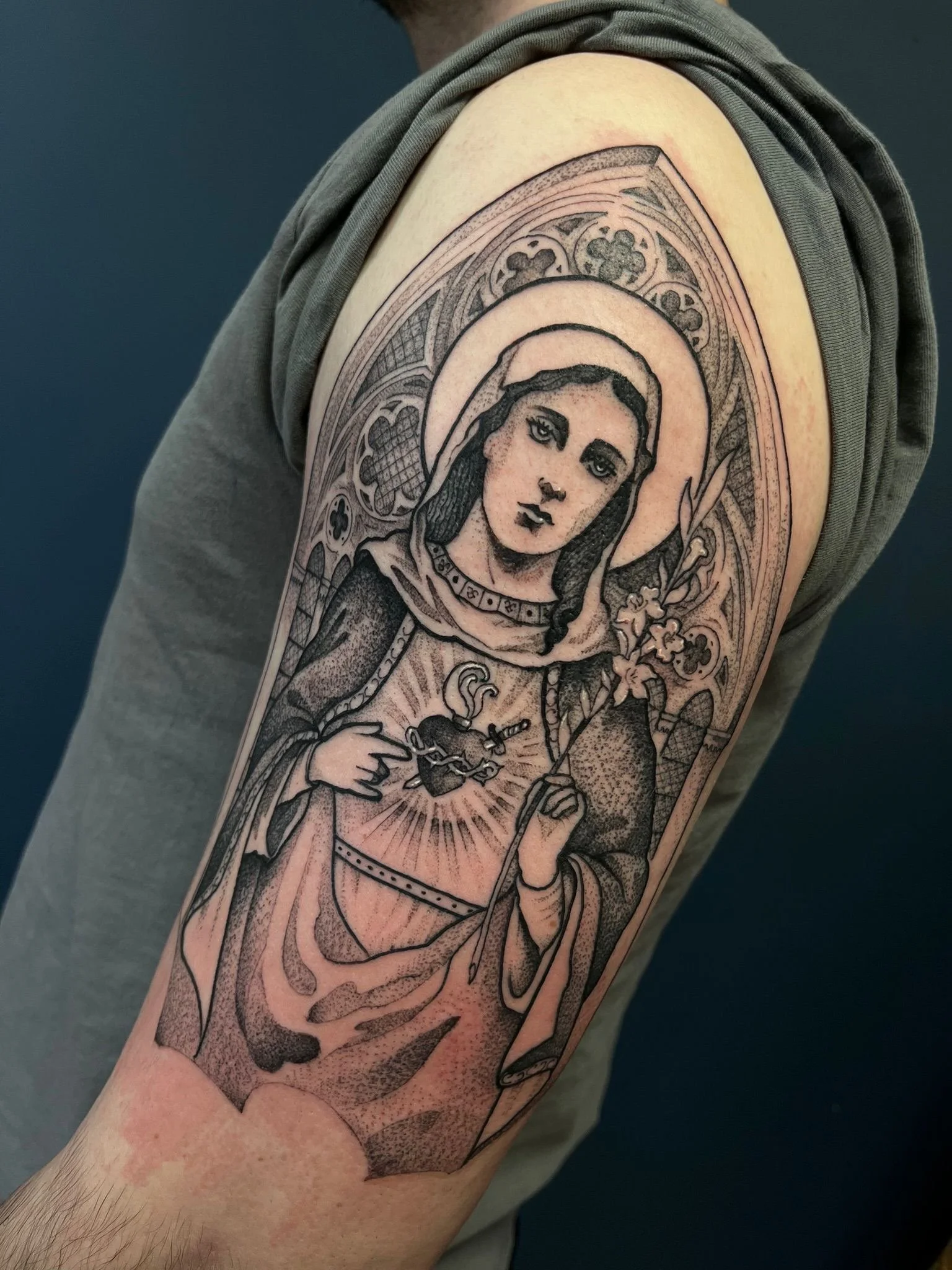 Black and gray tattoo of a religious figure, possibly a saint or the Virgin Mary, holding a lily in one hand and a heart emitting rays of light, with intricate patterns in the background.