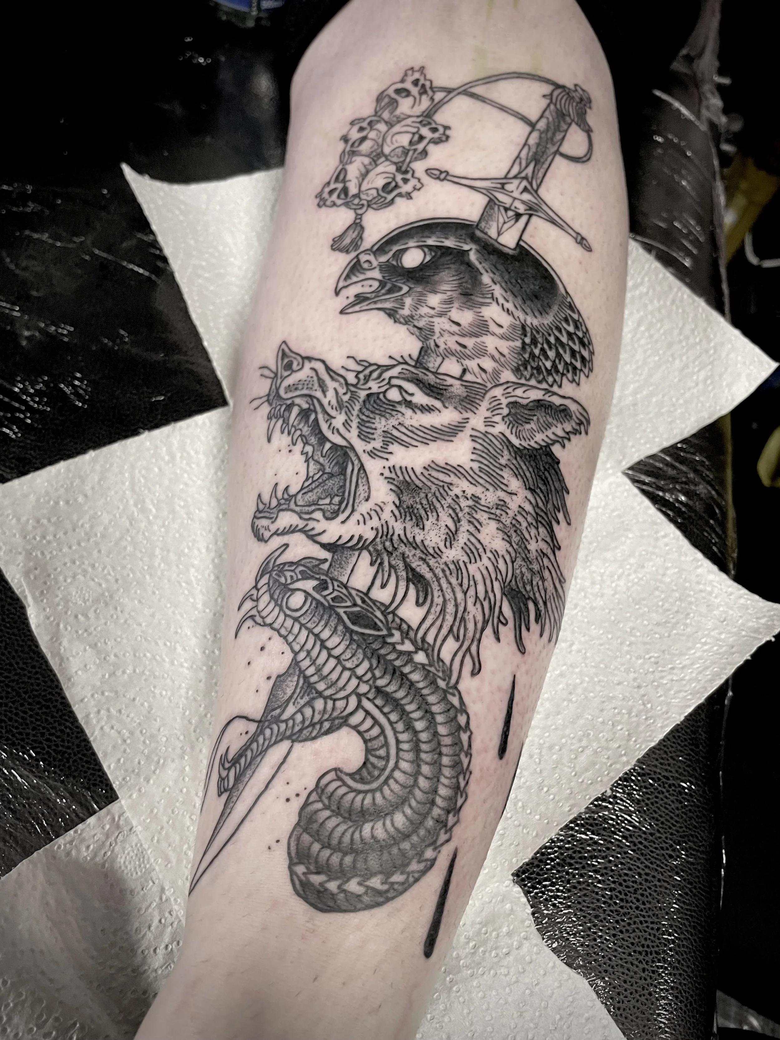 Black ink tattoo on a person's shin featuring a falcon, a roaring wolf, a coiled snake, and a dagger.
