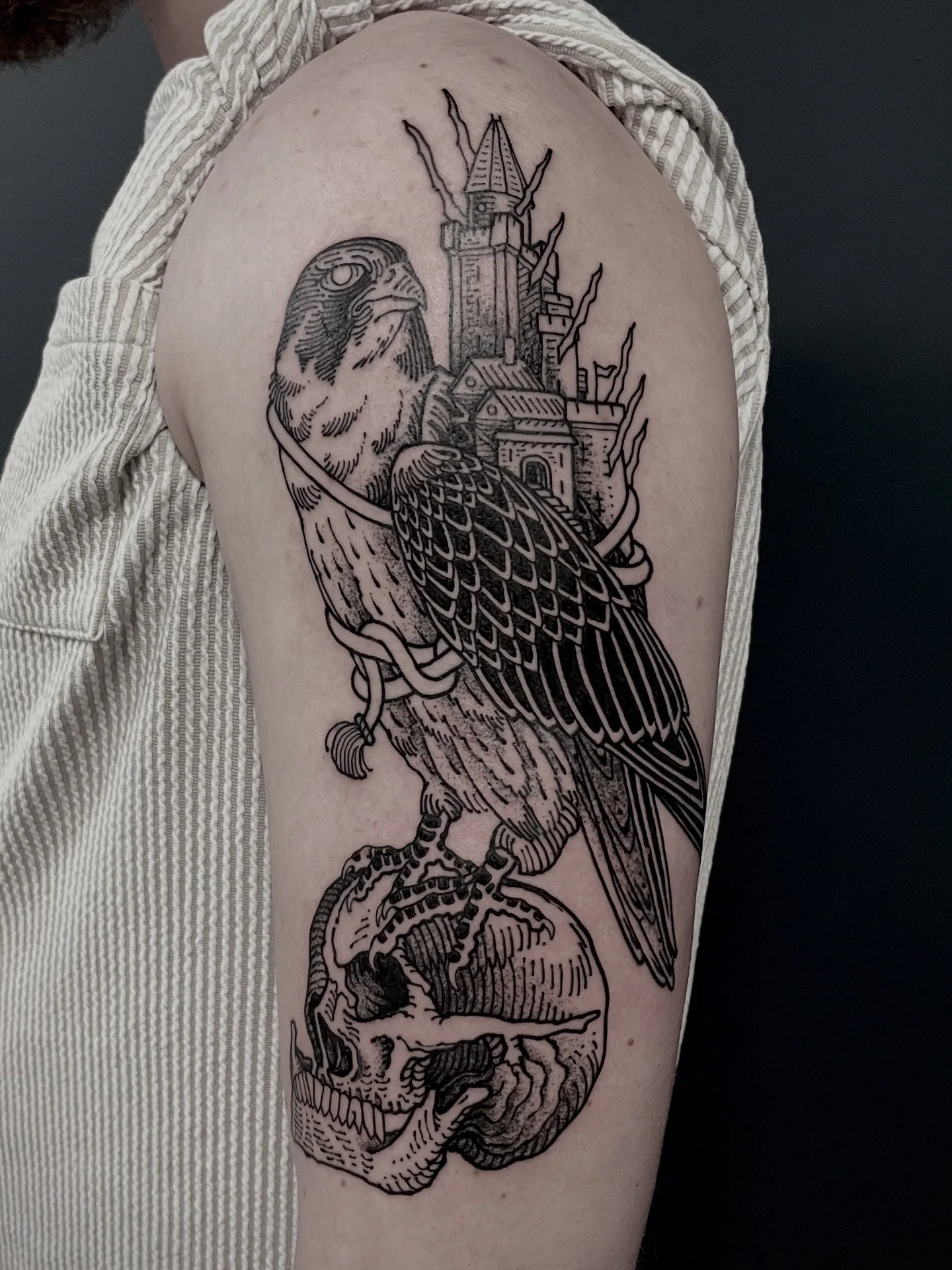 A black outline tattoo of a falcon with detailed feathers perched on a human skull with a cityscape and a tall tower in the background.