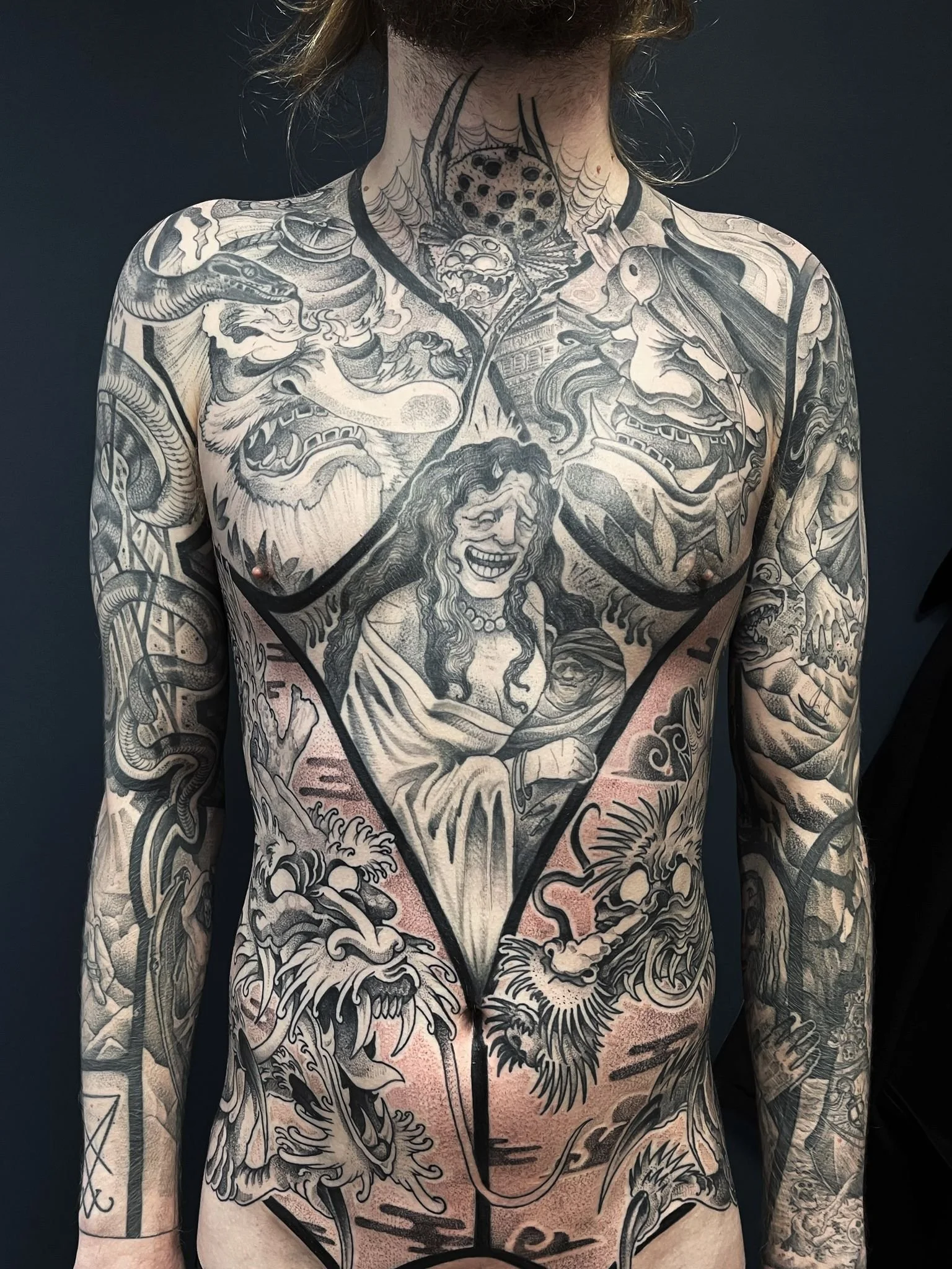 Extensive black and gray tattoo covering a man's chest, torso, and arms, featuring various mythological, traditional, and fantastical designs including a dragon, lions, a woman with a wide smile, a spider, and other intricate patterns.
