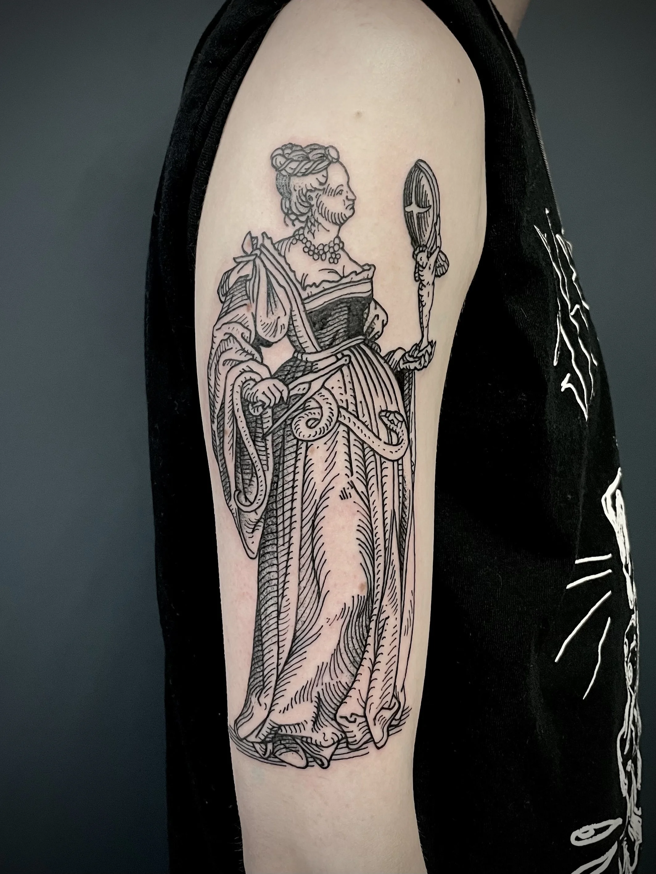 Black ink tattoo of a medieval woman holding a mirror in her right hand, dressed in a detailed gown with a snake at her waist, on person's upper arm.