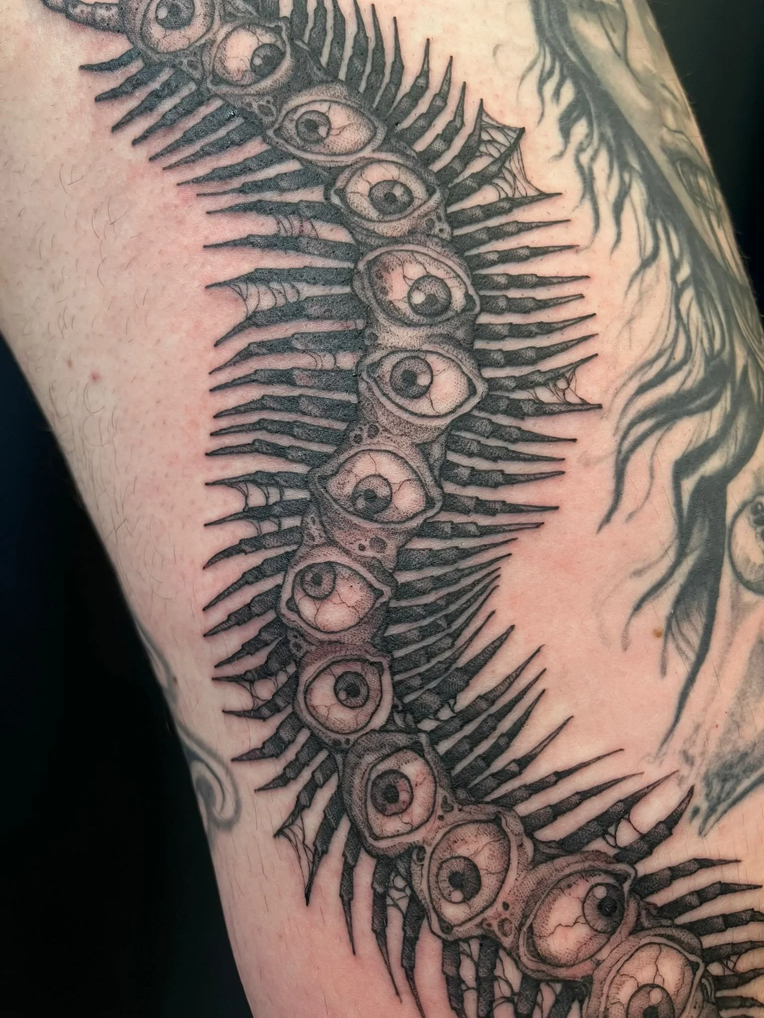 Black and gray tattoo of a row of eyes and spikes, arranged vertically on a person's arm.