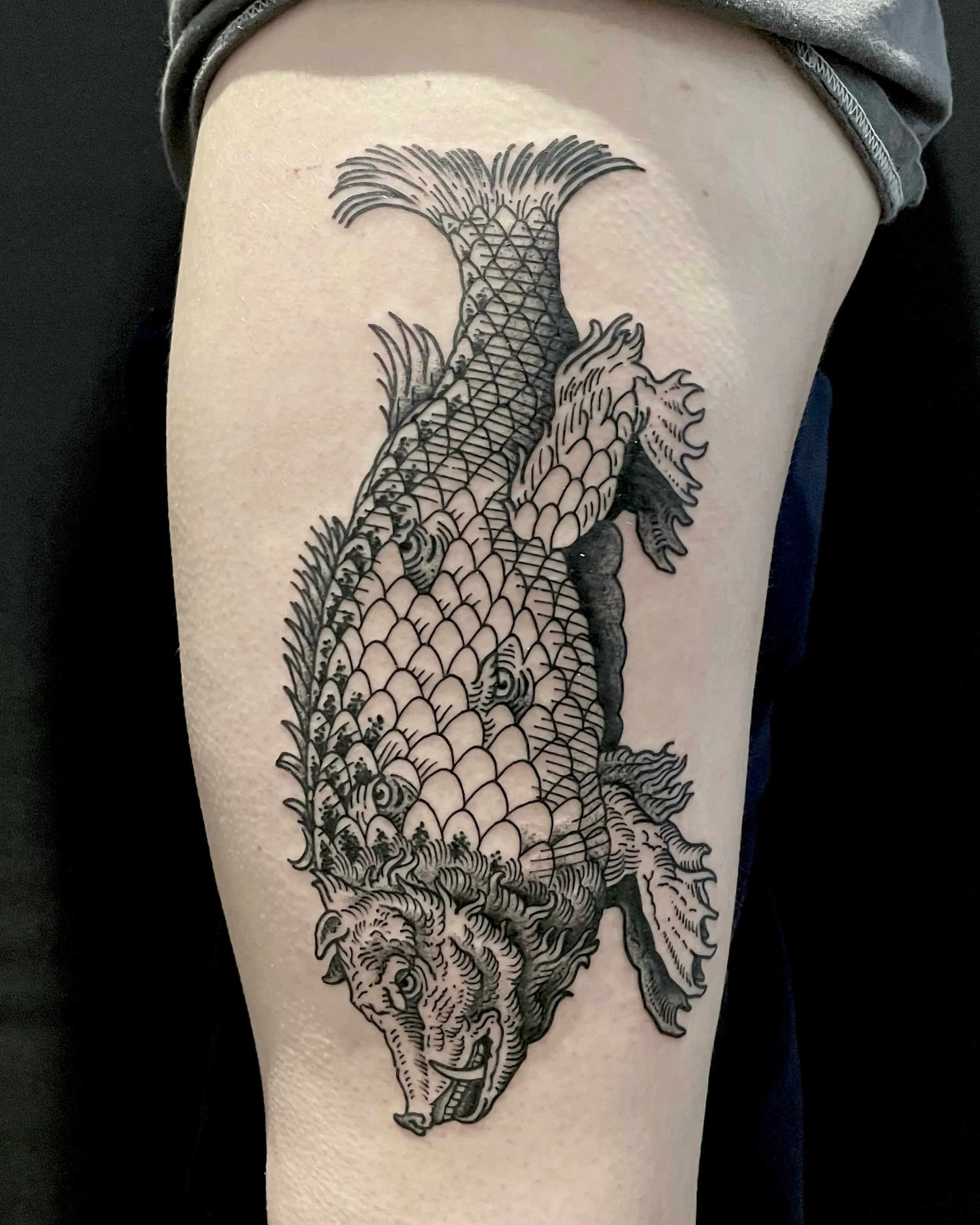 Line art style tattoo of a fish monster on person’s thigh.