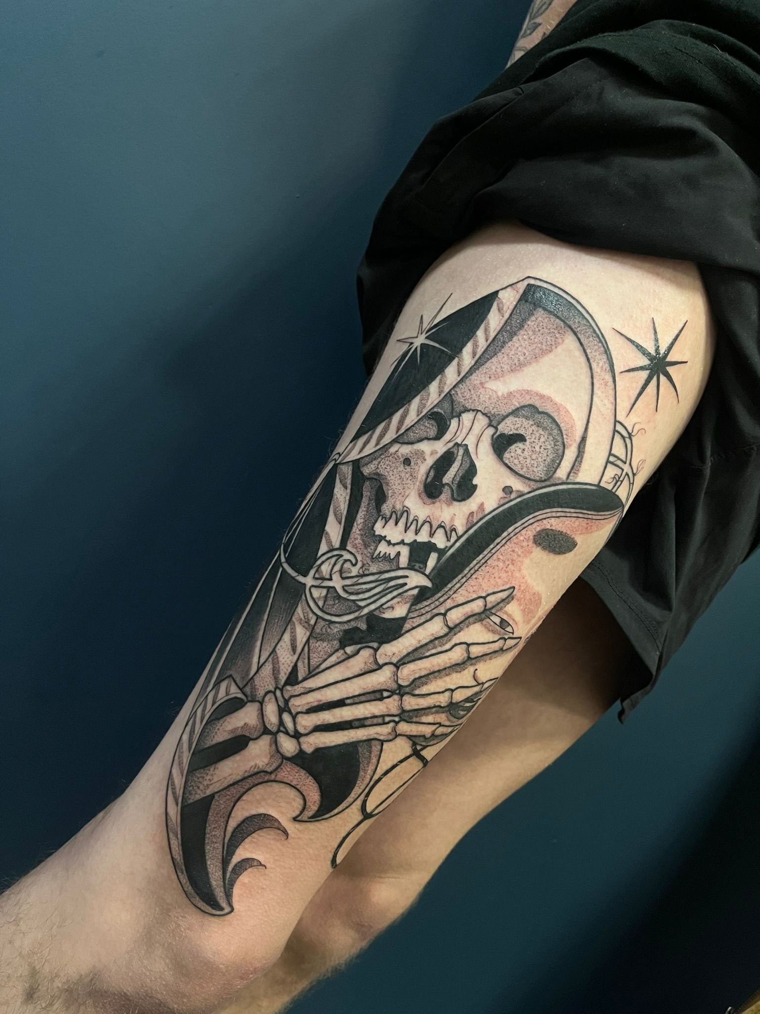 Tattoo of a skeleton with a skull, holding a mask, with stars around it, on a person's thigh.