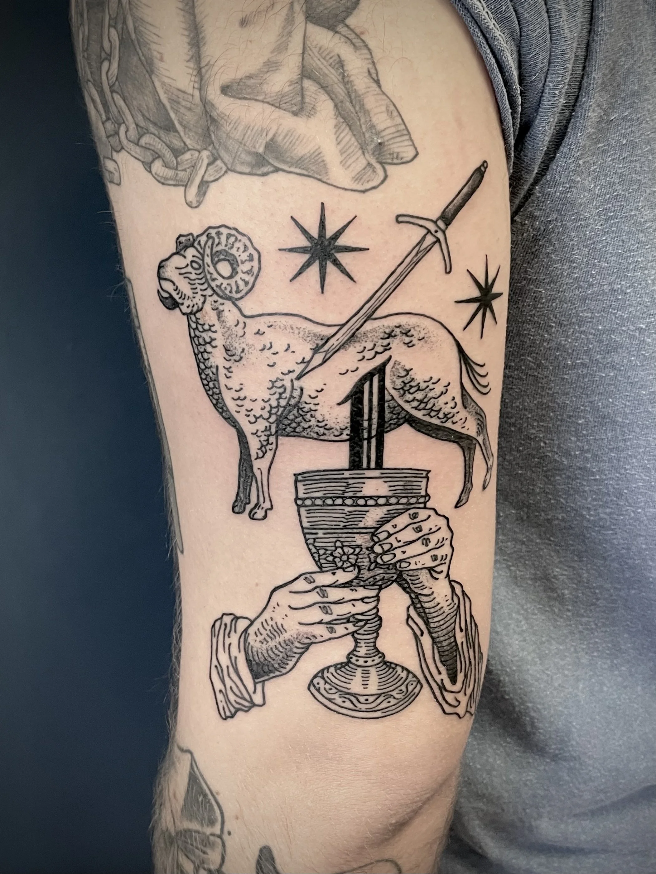 Black line tattoo depicting a lamb, a sword stabbed into a horn-shaped vessel, a human hand holding the vessel, and three stars around.