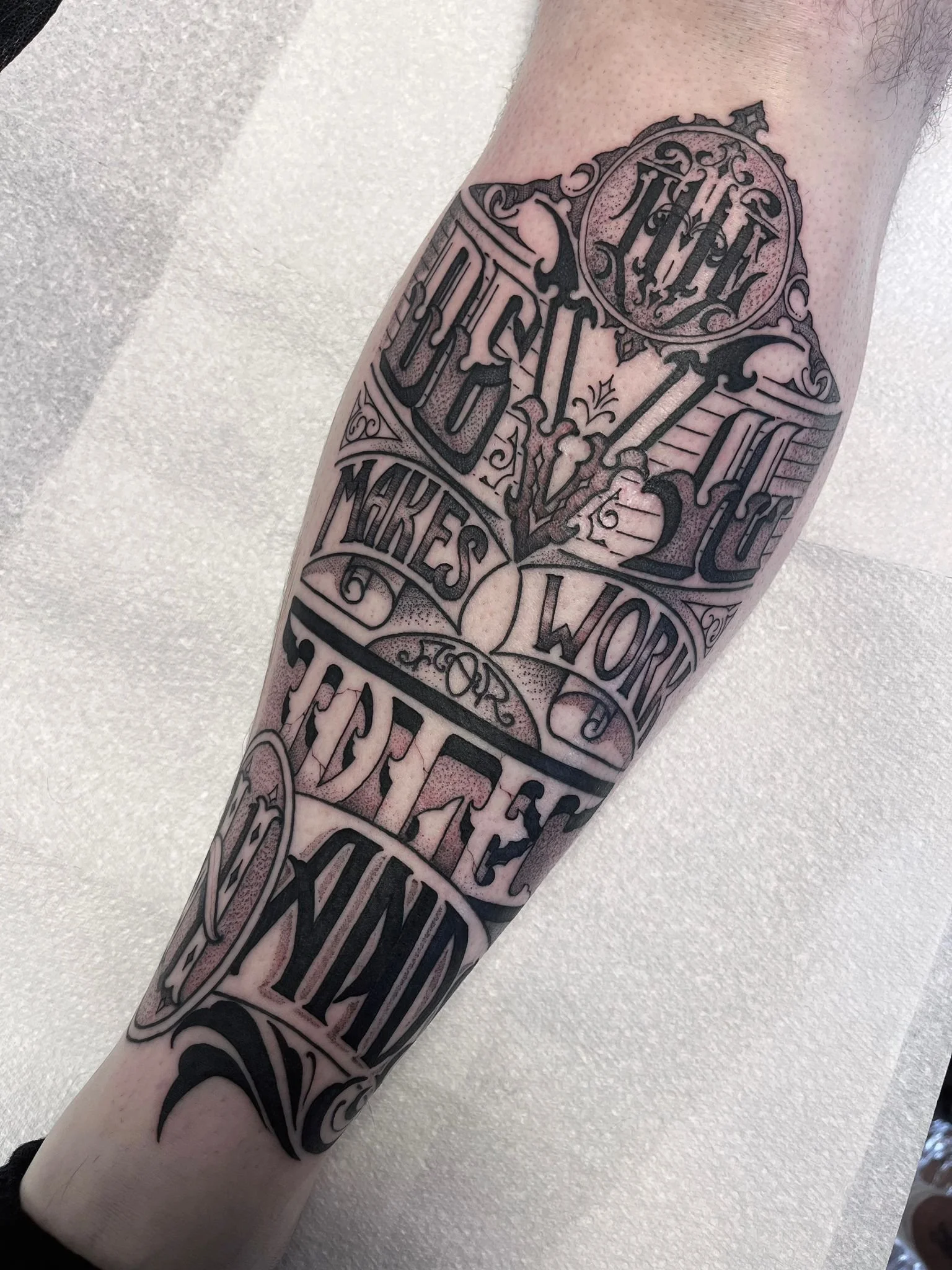Black and gray tattoo on a person's forearm with ornate lettering and decorative elements, featuring a quote about making words beautiful.