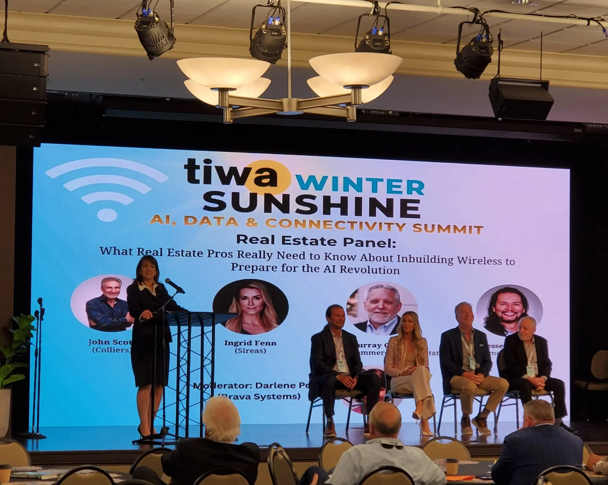 Panel at the TIWA Winter Sunshine AI, Data & Connectivity Summit with five speakers, three men and two women, sitting on stage and one woman standing at a podium. The large screen behind displays the event name, theme, and a real estate panel discussion about wireless inbuilding for the AI revolution.