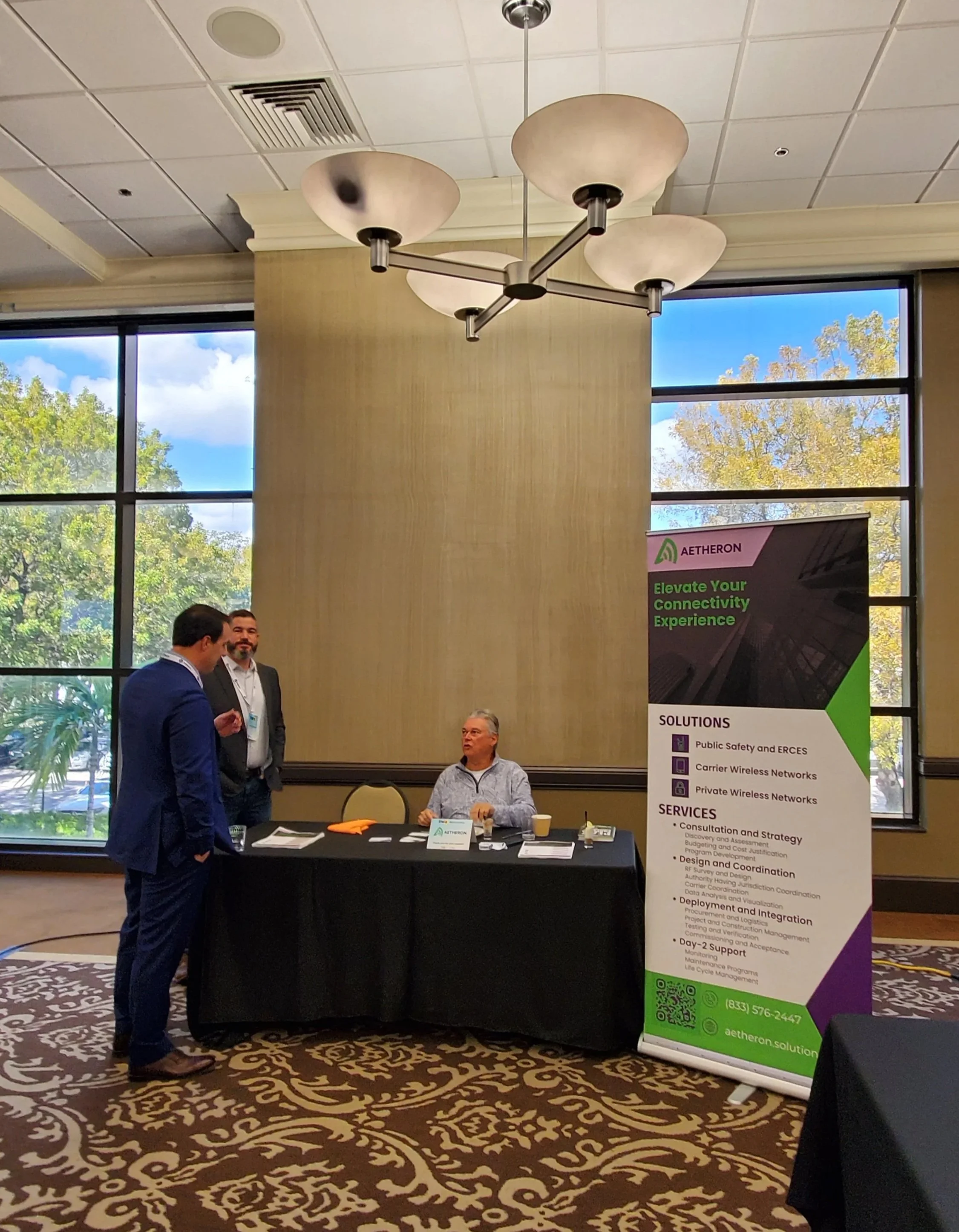 A man in a blue suit is talking to two men behind a booth at a trade show or conference. The booth has a black tablecloth, with brochures and a nameplate. A large banner on the right promotes Aetheron solutions for wireless networks and public safety. The room has large windows showing trees and a blue sky outside, with a patterned carpet and a modern chandelier overhead.