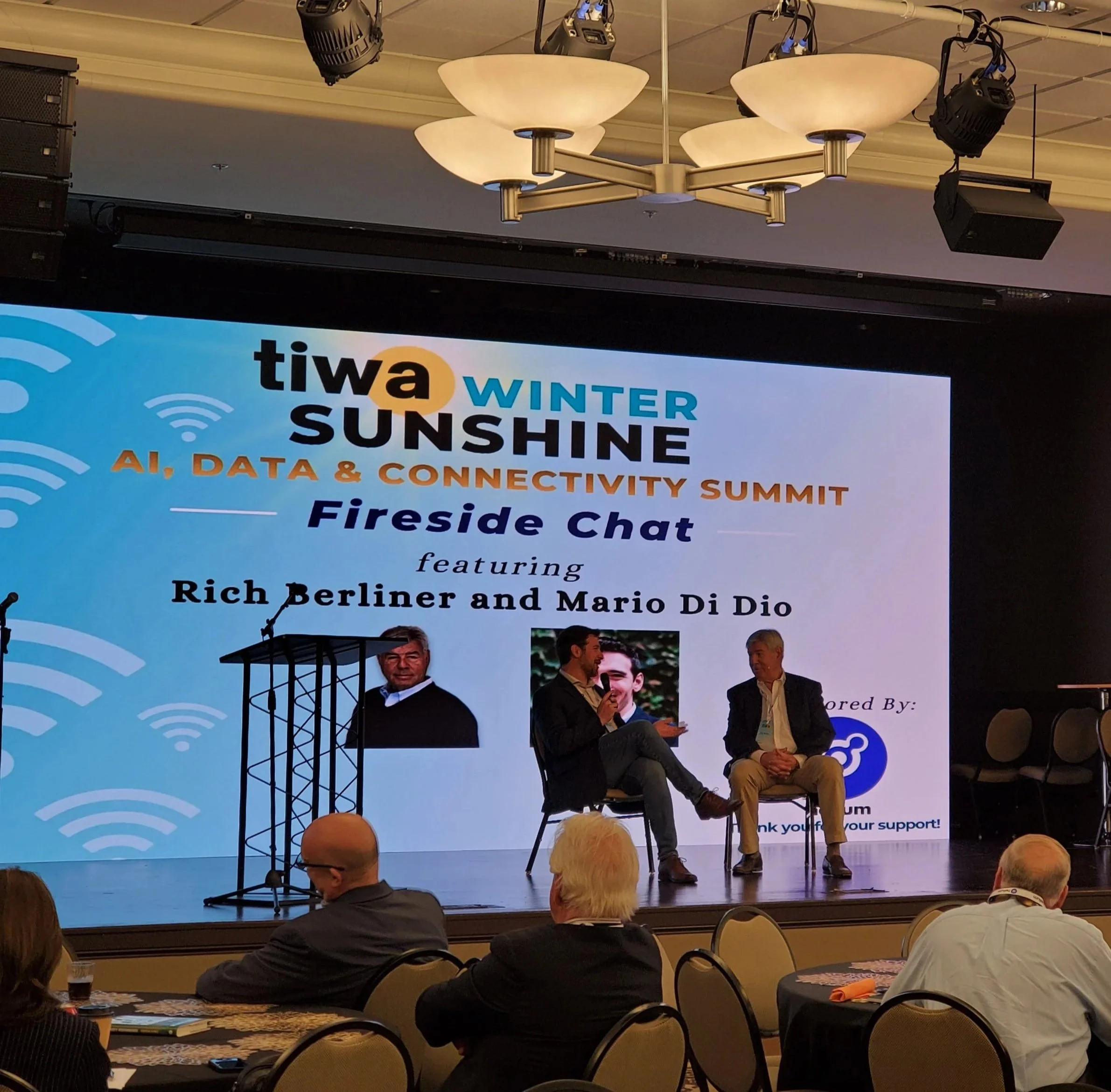 Two men seated on a stage engaged in a conversation during a panel discussion at the TIWA Winter Sunshine AI, Data & Connectivity Summit. A large screen behind them displays the event's name and speakers, with the audience seated at round tables.