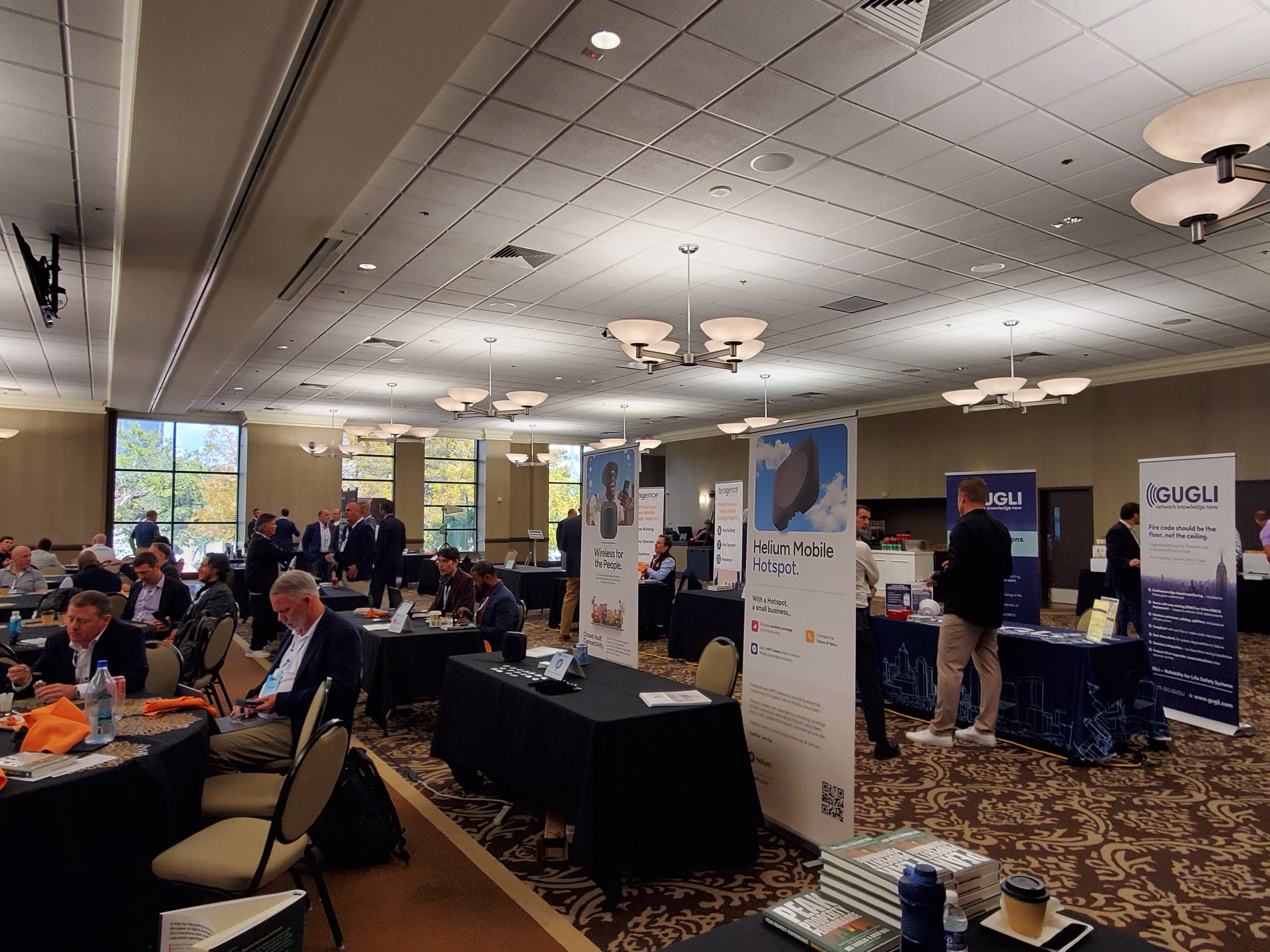 A conference room filled with attendees sitting at tables and walking around, with banners and display tables set up for a tech event.