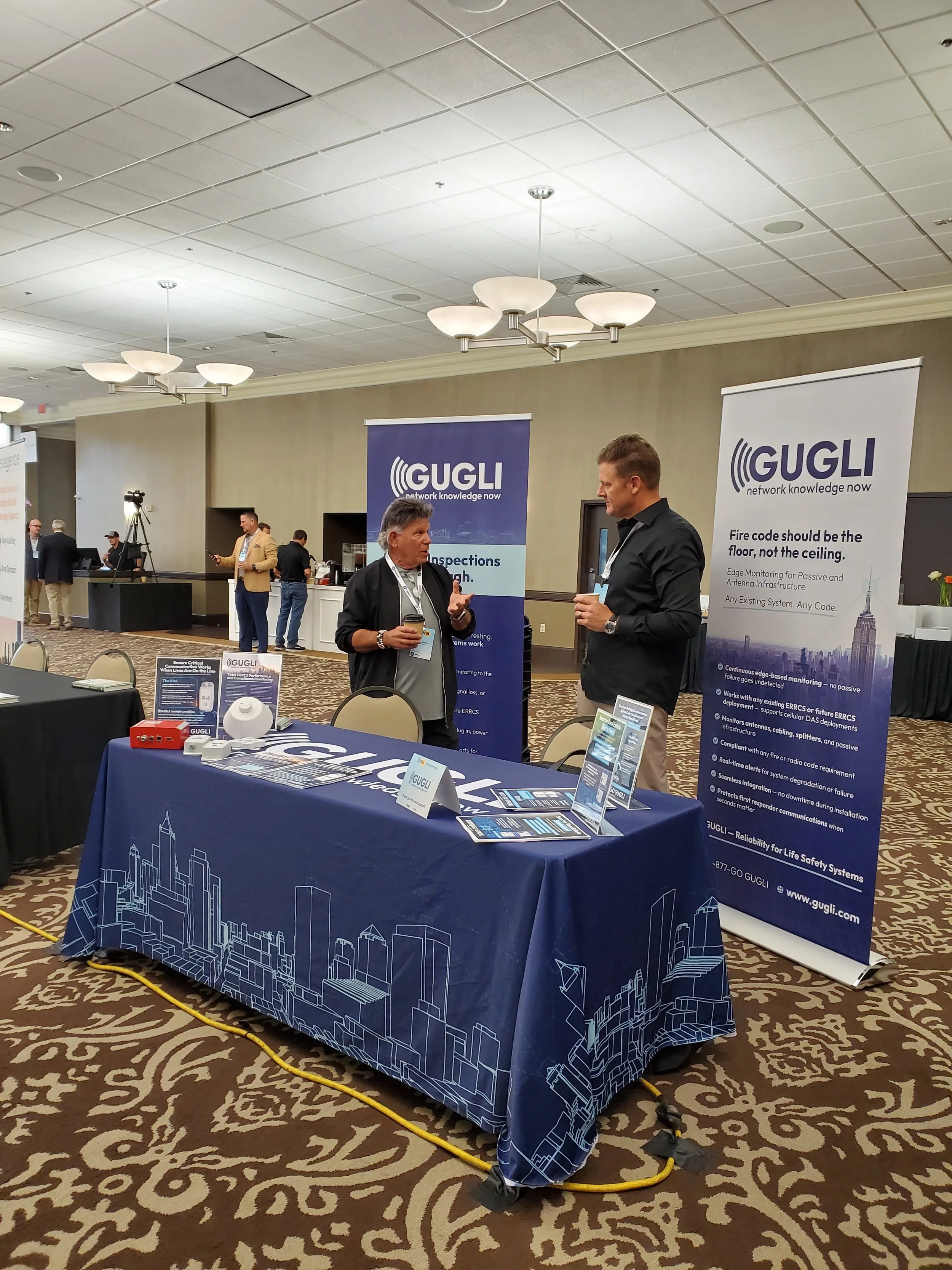 GUGLI conference booth with two men talking, informational banners, brochures, and a tablecloth featuring a city skyline design.