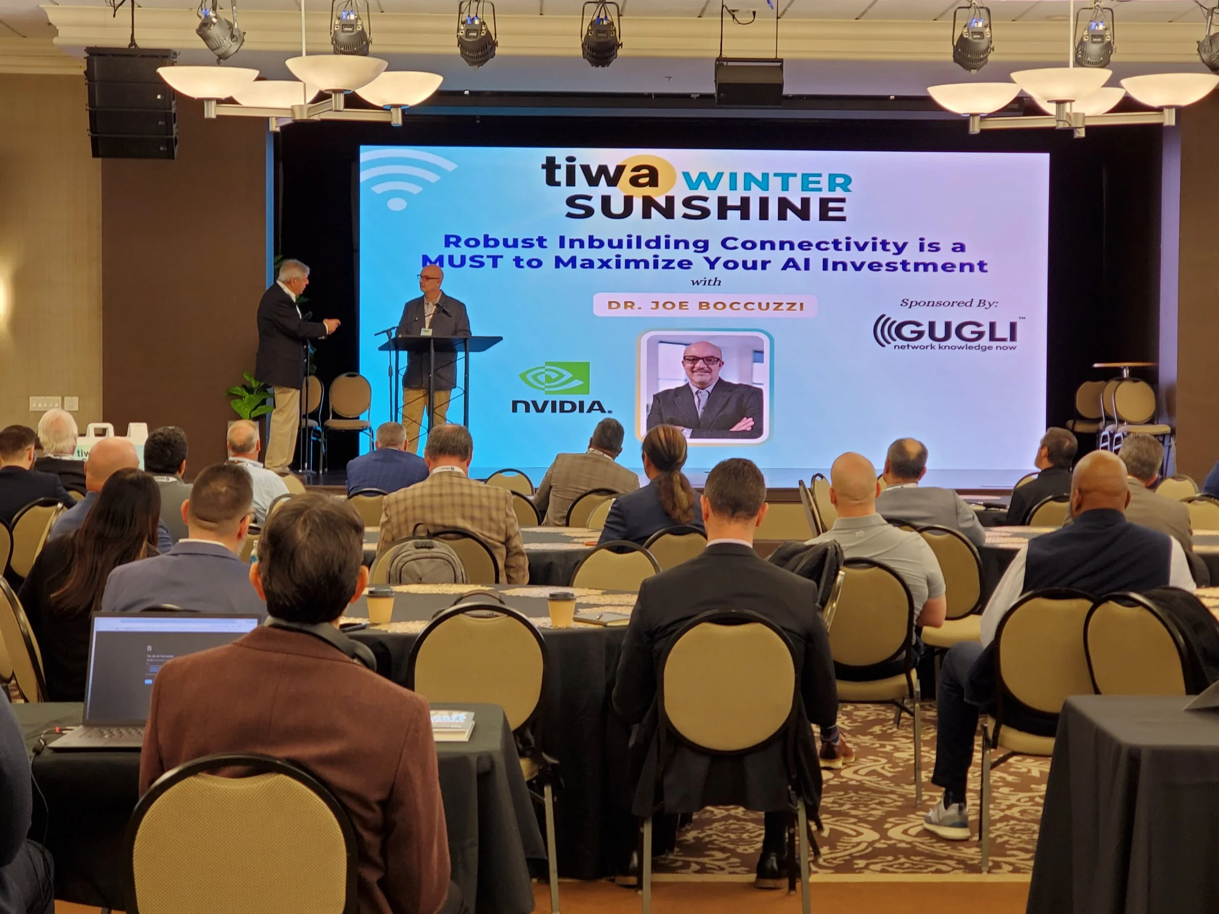 A conference room with attendees seated and two speakers on stage. A large screen behind displays a presentation titled 'tiwa WINTER SUNSHINE' with the subtitle 'Robust Inbuilding Connectivity is a MUST to Maximize Your AI Investment' and a photo of Dr. Joe Boccuzi. The event is sponsored by GUGLI.
