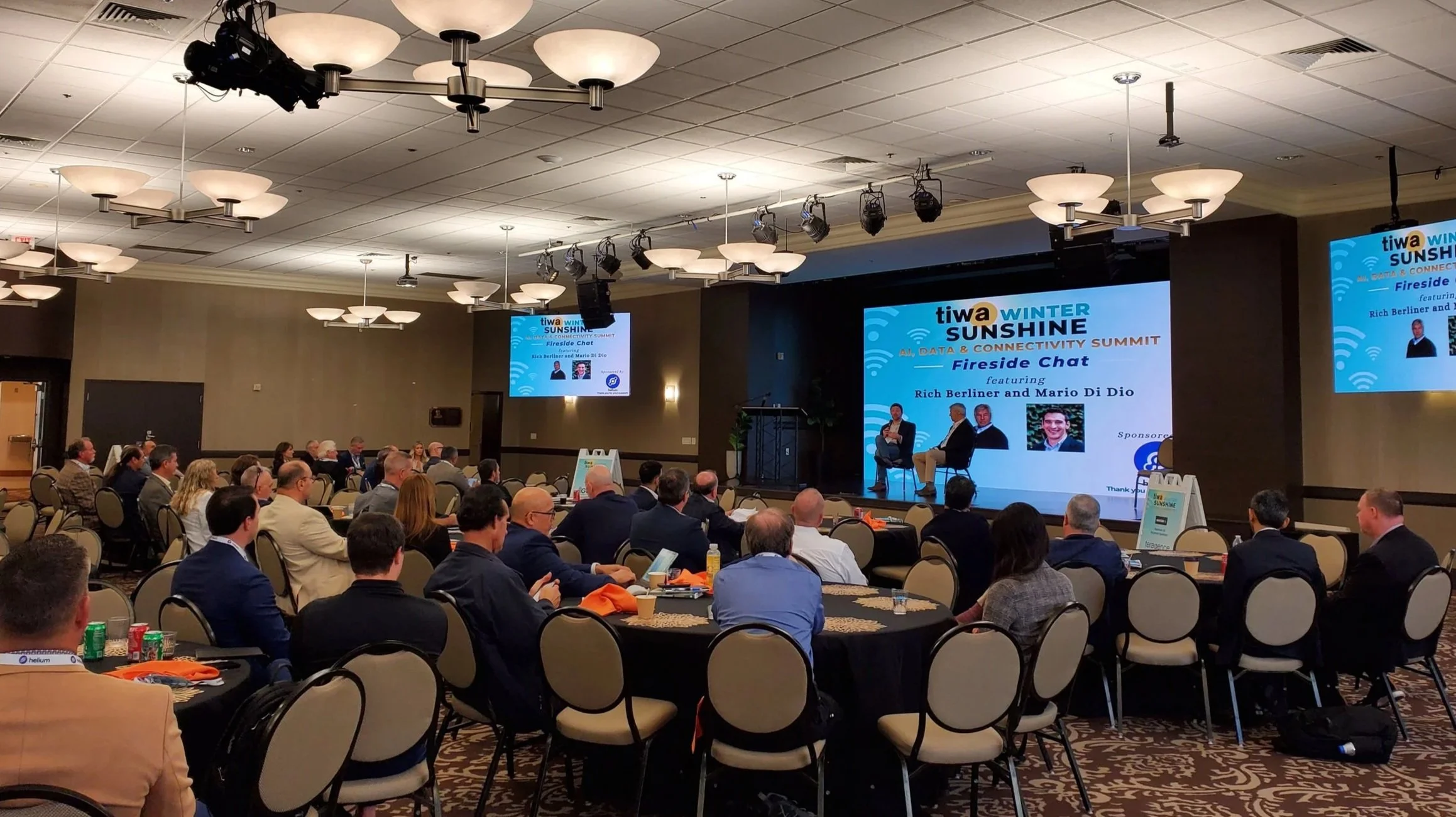 A conference room filled with seated attendees listening to a panel on stage at the fictional event 'tiwa WINTER SUNSHINE All, Data & Connectivity Summit'. The stage features a large screen with the event title and panelist names, and three panelists are seated and speaking.