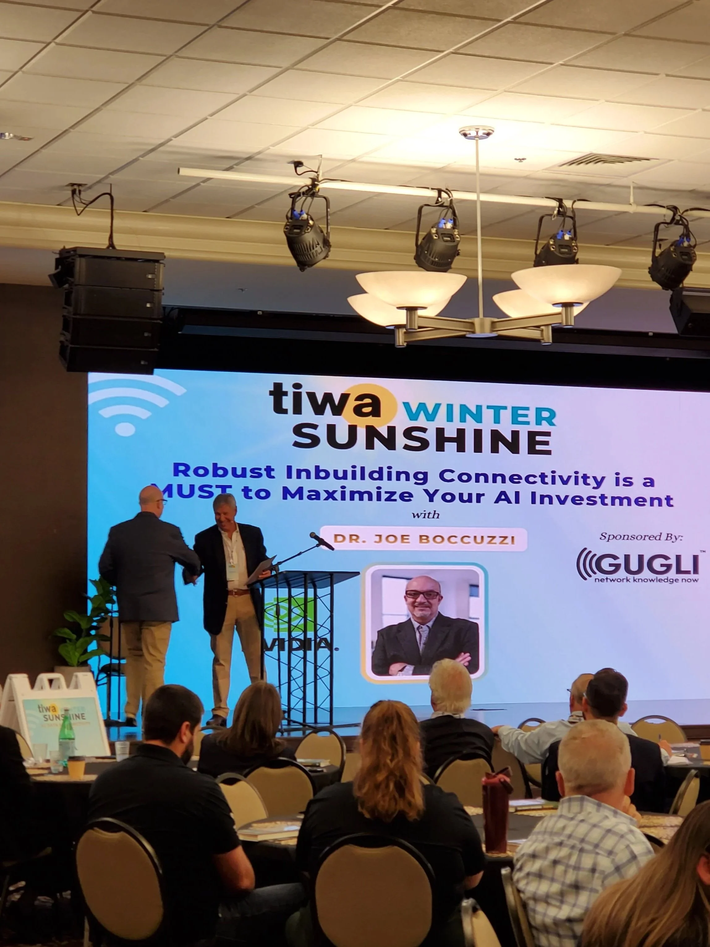 A conference room with a stage displaying a presentation slide about AI investment, featuring a photo of Dr. Joe Boccuzzi. Two men on stage shake hands in front of seated audience members, with a large screen behind them showing the event's details.